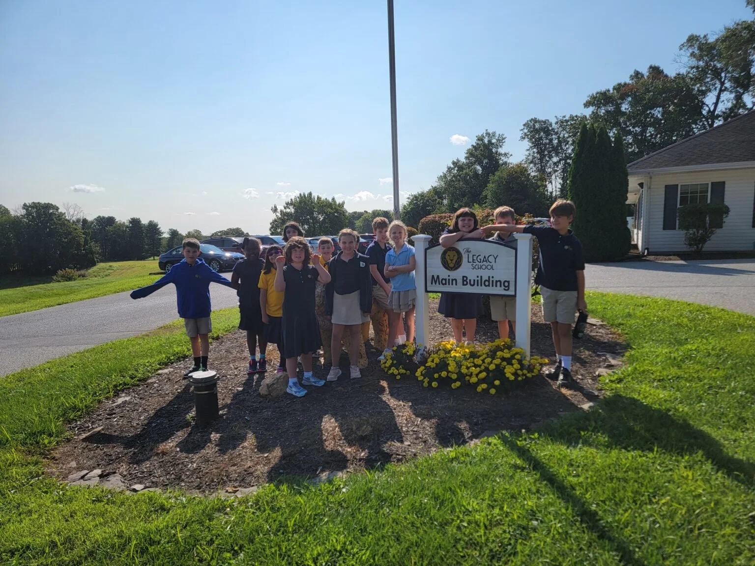 Group of children standing outdoors near school sign that reads 'Legacy School Main Building', with a grassy area, parked cars, and a white building in the background on a sunny day.
