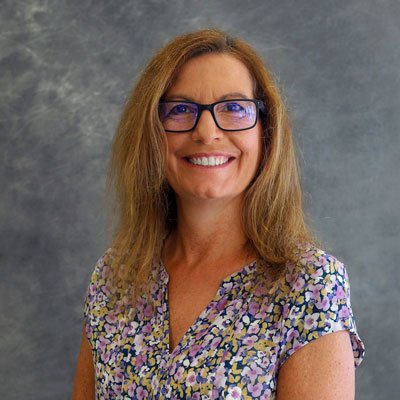 A woman with shoulder-length light brown hair, wearing glasses and a floral blouse, smiling in front of a gray background.