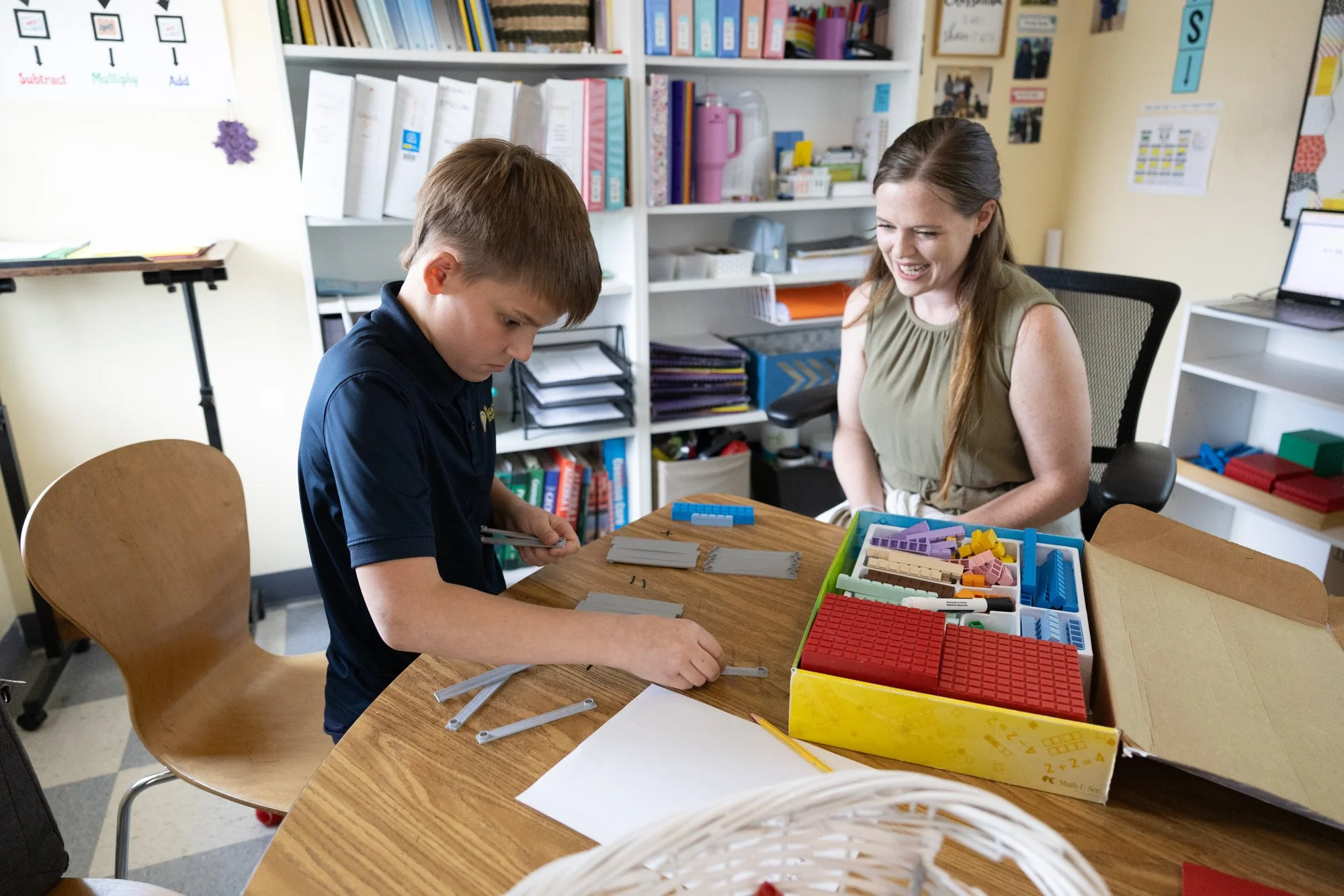 A boy and a woman sit at a desk, playing with educational toys. The boy is looking down, focused on the toys, while the woman smiles and looks at him. Behind them are shelves filled with books, binders, and office supplies, with a whiteboard on the wall.