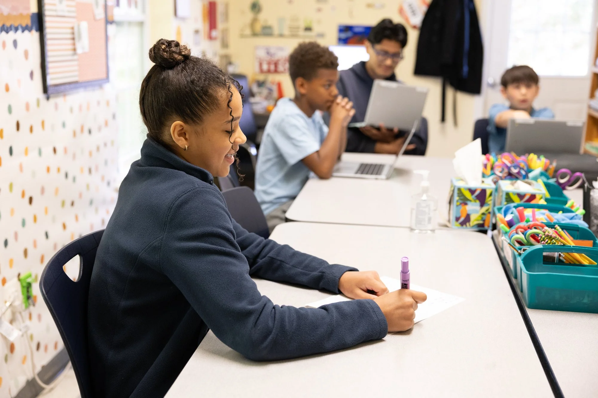 A young girl sitting at a table with a purple marker in her hand, smiling as she writes on paper, with colorful school supplies in front of her. In the background, three children and an adult with a laptop are engaged in a classroom setting.
