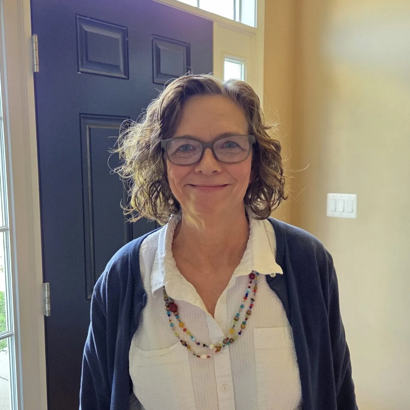 A woman with curly brown hair and glasses smiling at the camera, standing inside a house near a blue door and yellow wall, dressed in a white shirt and colorful beaded necklace.