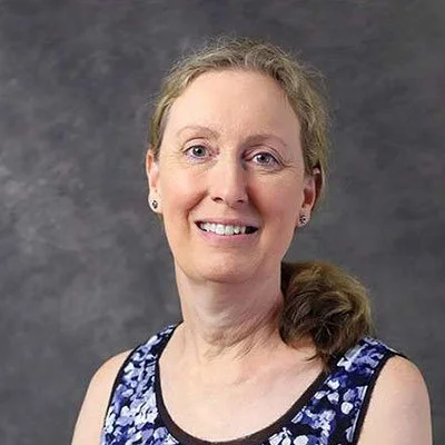 A woman with light skin and blonde hair smiling at the camera, wearing a sleeveless blue and white patterned top against a dark gray background.