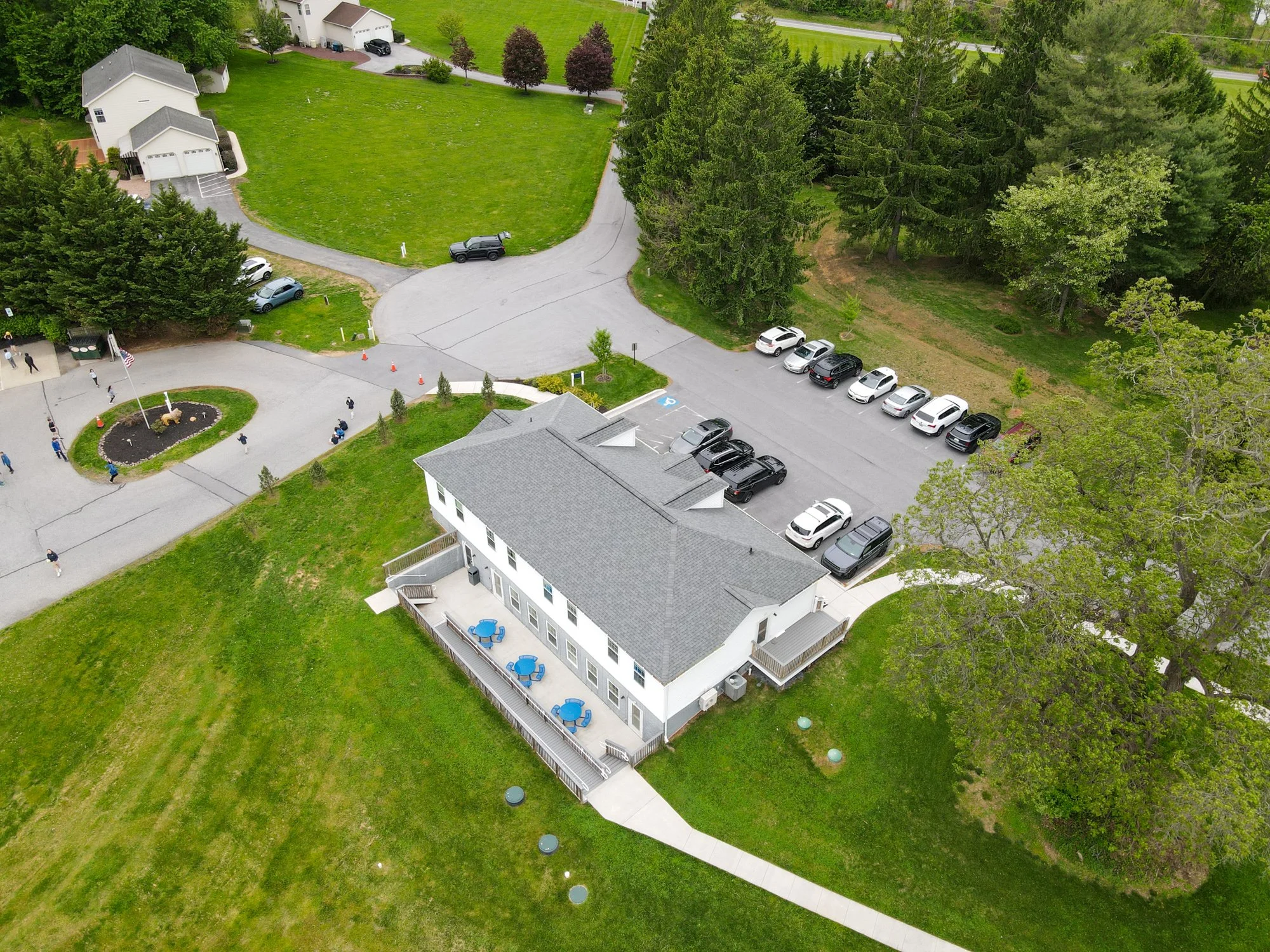 An aerial view of a neighborhood showing a white residential building with a balcony and a backyard patio, a parking lot with several cars, a green lawn, numerous trees, and a small landscaped area with a flag. There are pedestrians walking along the sidewalk and small orange cones blocking part of the driveway.