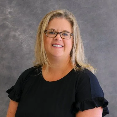 A woman with blonde hair and glasses smiling, wearing a black shirt with ruffled sleeves, standing in front of a gray textured background.