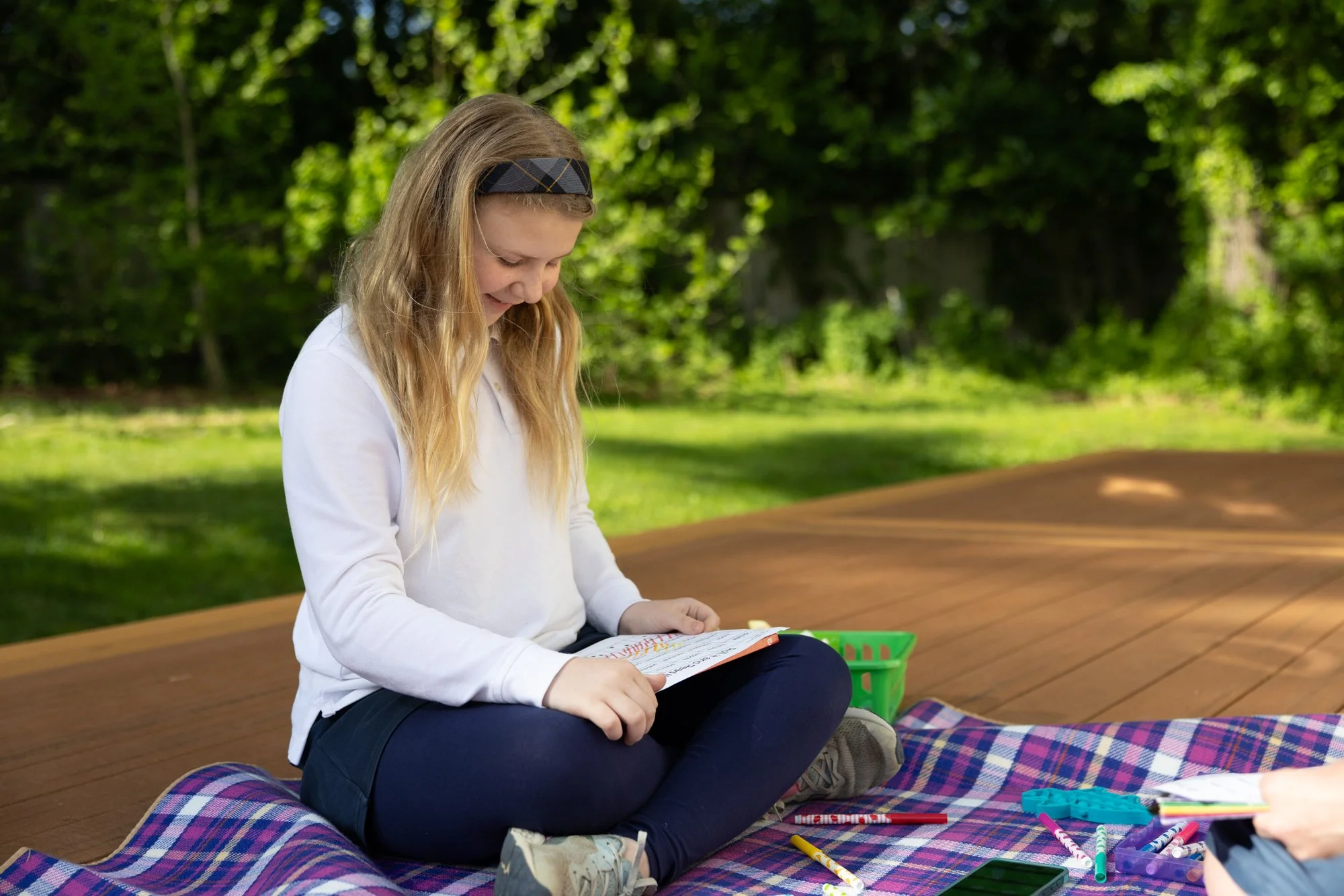 A young girl with long blonde hair and a black headband, sitting outdoors on a picnic blanket, looking down at a notebook, surrounded by colored markers and pens, with trees and greenery in the background.