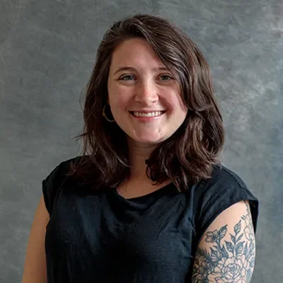 A smiling woman with shoulder-length brown hair, wearing a black t-shirt and hoop earrings, standing against a gray background, showing her tattooed left arm.