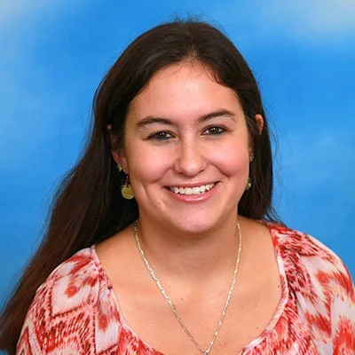 A young woman with long dark hair smiling in front of a blue background, wearing a red and white patterned top and gold earrings.