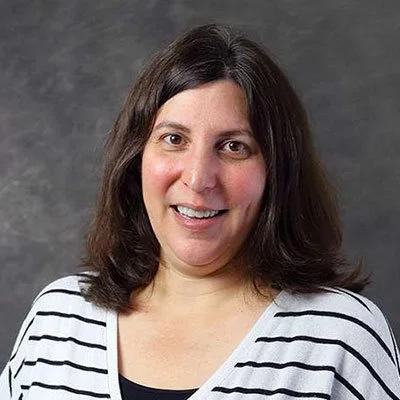 A woman with shoulder-length dark hair smiling at the camera, wearing a white and black striped shirt, in front of a gray background.