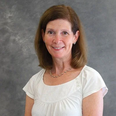 A woman with shoulder-length brown hair, smiling, wearing a white blouse and a necklace, standing against a gray background.