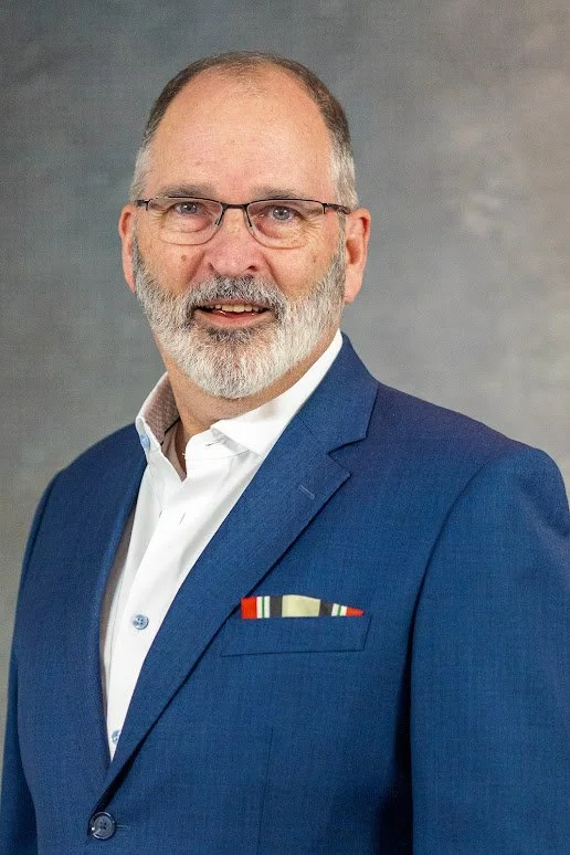 A man with glasses, a beard, and short hair, wearing a blue suit and white shirt, posing against a gray background.