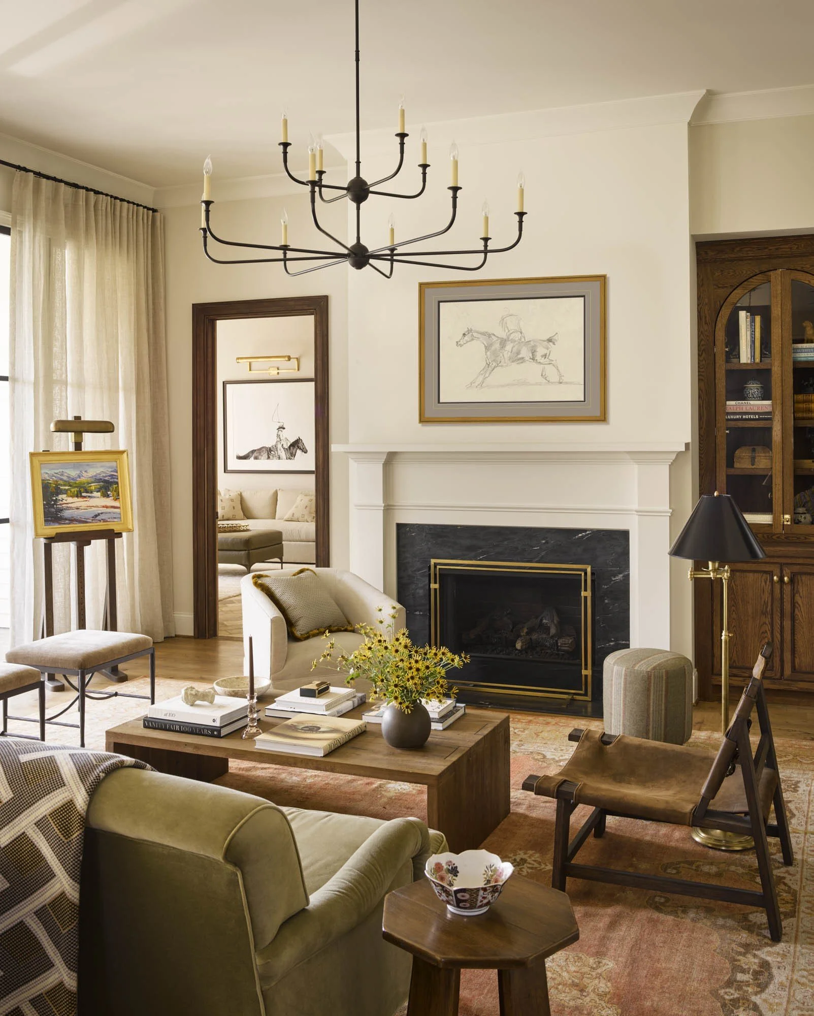 Living room with a beige sofa, patterned pillows, a wooden coffee table with books and a vase of yellow flowers, a black fireplace, framed artwork, a black chandelier, a window with curtains, and various chairs and side tables.