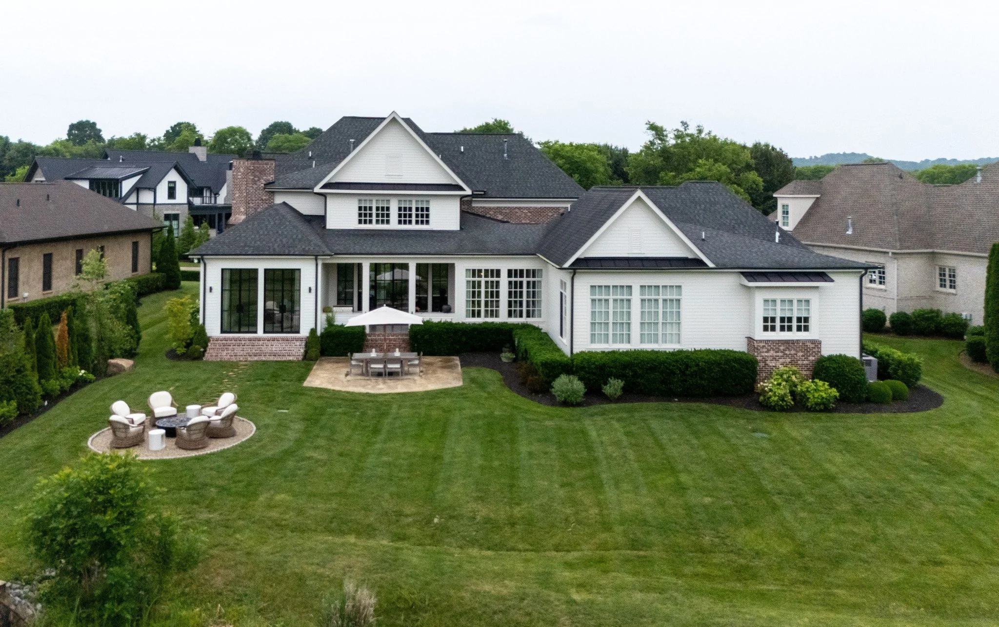 A large backyard with a green lawn, outdoor furniture including chairs and a white umbrella, and a white house with multiple windows and a patio. Neighboring houses are visible in the background with trees and a cloudy sky.