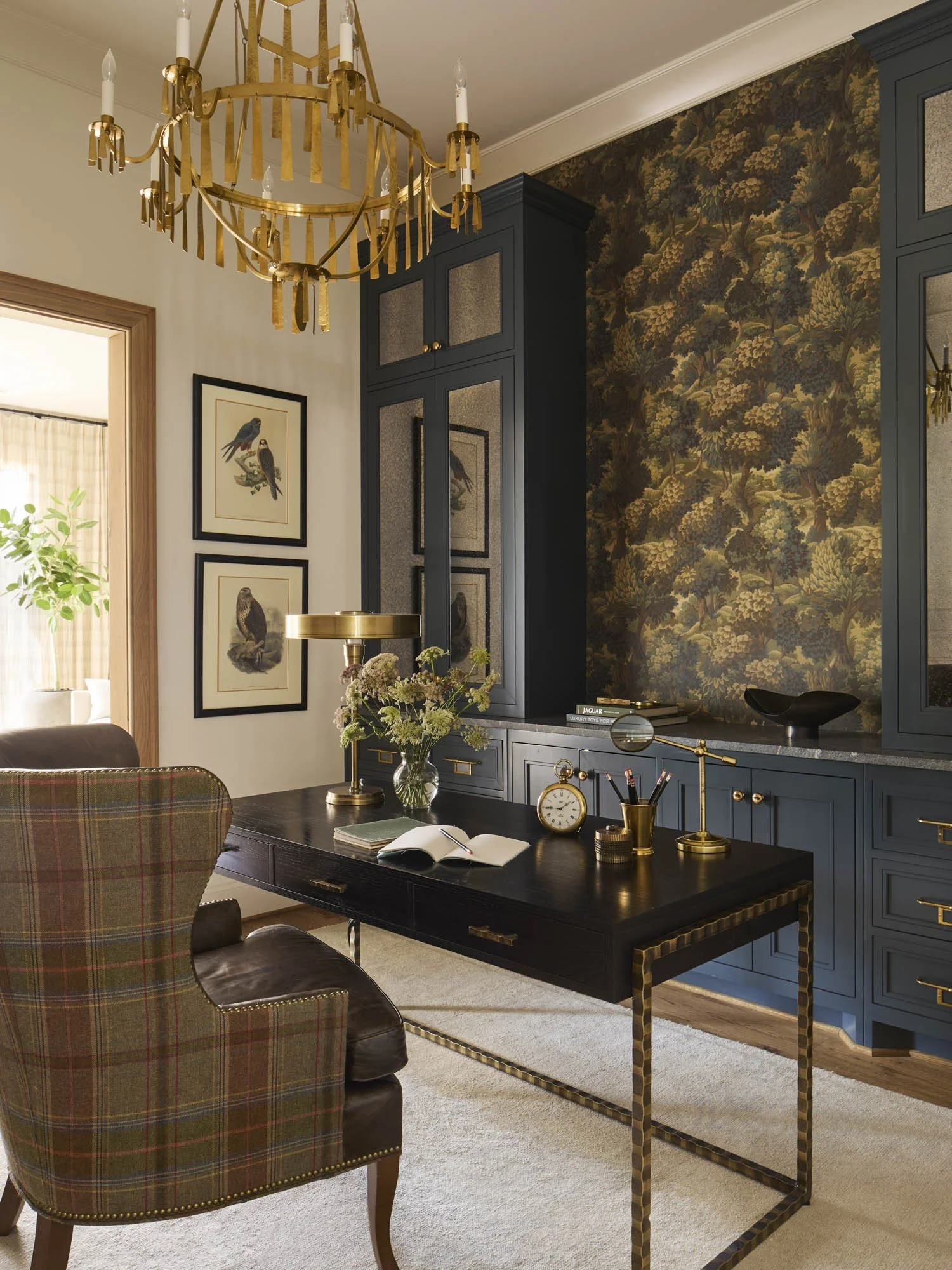 A living room corner with a black cabinet, framed bird art on the wall, a gold chandelier, a dark wooden desk with an open book, a brass clock, and a vase of flowers. A plaid upholstered chair is in the foreground.