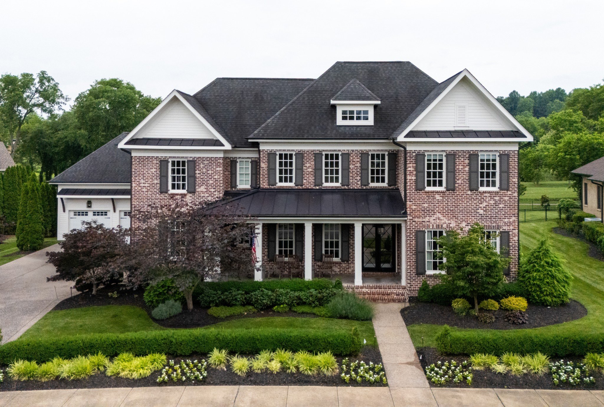 Front view of a two-story house with a brick facade, black shutters, and a dark roof, surrounded by a landscaped yard with green grass, shrubs, and trees.