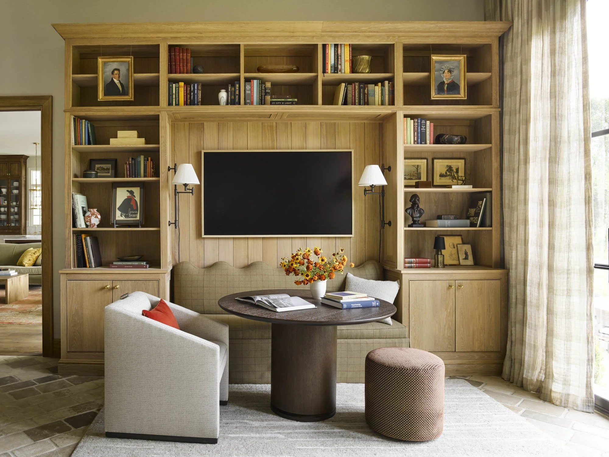 A living room with a wooden bookshelf wall, a flat-screen television, a loveseat with cushions, two side lamps, a round table with a book and flowers, and additional seating with a patterned ottoman.