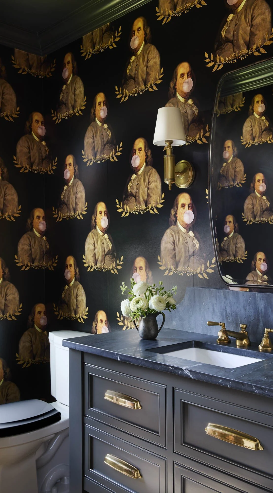 Bathroom with wallpaper featuring portraits of Benjamin Franklin wearing face masks, a white flowers in a black vase on the dark gray countertop, a white toilet, a wall-mounted lamp, and a mirror reflecting the wallpaper.
