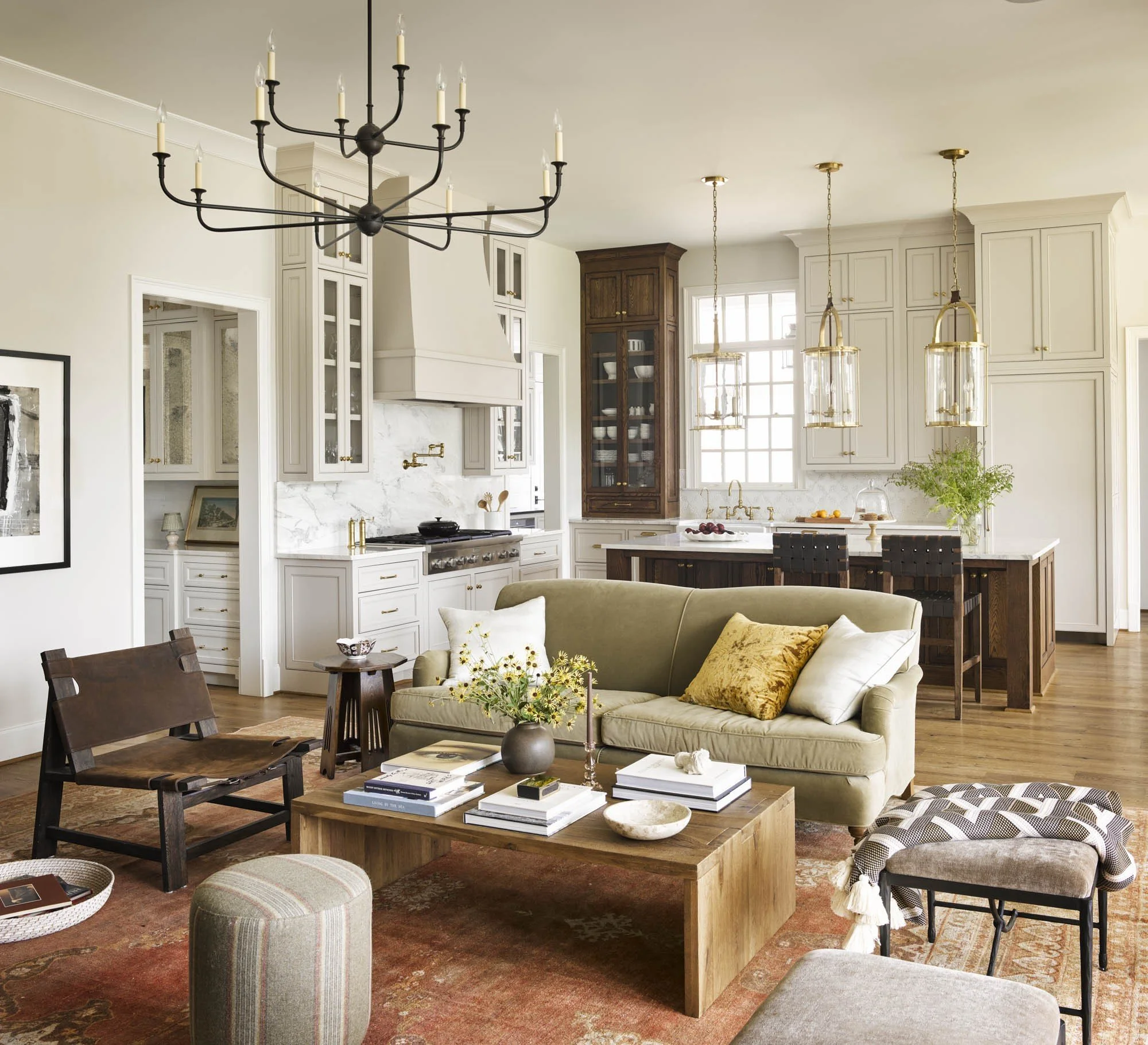 Living room and kitchen with modern and rustic decor, including a black chandelier, gold pendant lights, white cabinets, a marble backsplash, an olive-green sofa with yellow pillows, a wooden coffee table with books and decorative items, and a large 