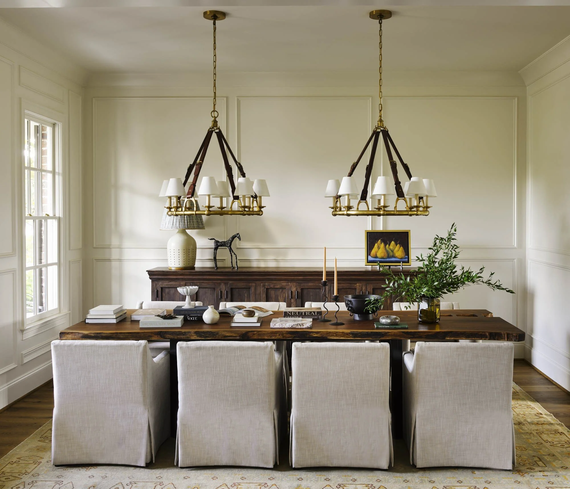 A dining room with cream-colored walls, two brass chandeliers hanging from the ceiling, a dark wooden dining table, eight white upholstered chairs, and decorative items like a white ceramic lamp, small sculptures, a framed painting of yellow fruit, a