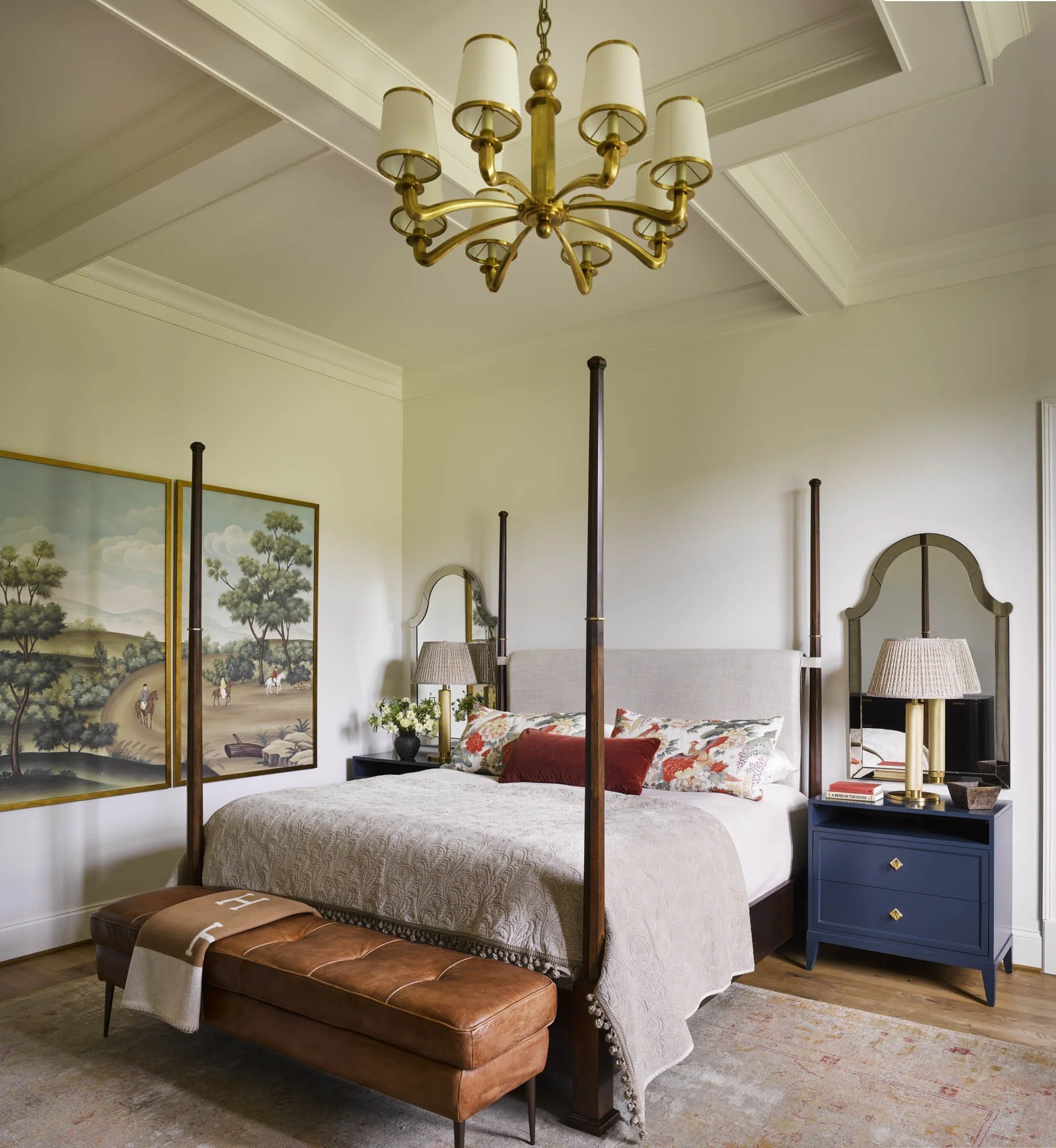 A bedroom with a four-poster bed, beige bedding, and a brown leather bench at the foot of the bed. There are two lamps on blue nightstands, a floral rug on a wooden floor, and a wall with paintings of trees. A gold chandelier hangs from the ceiling.