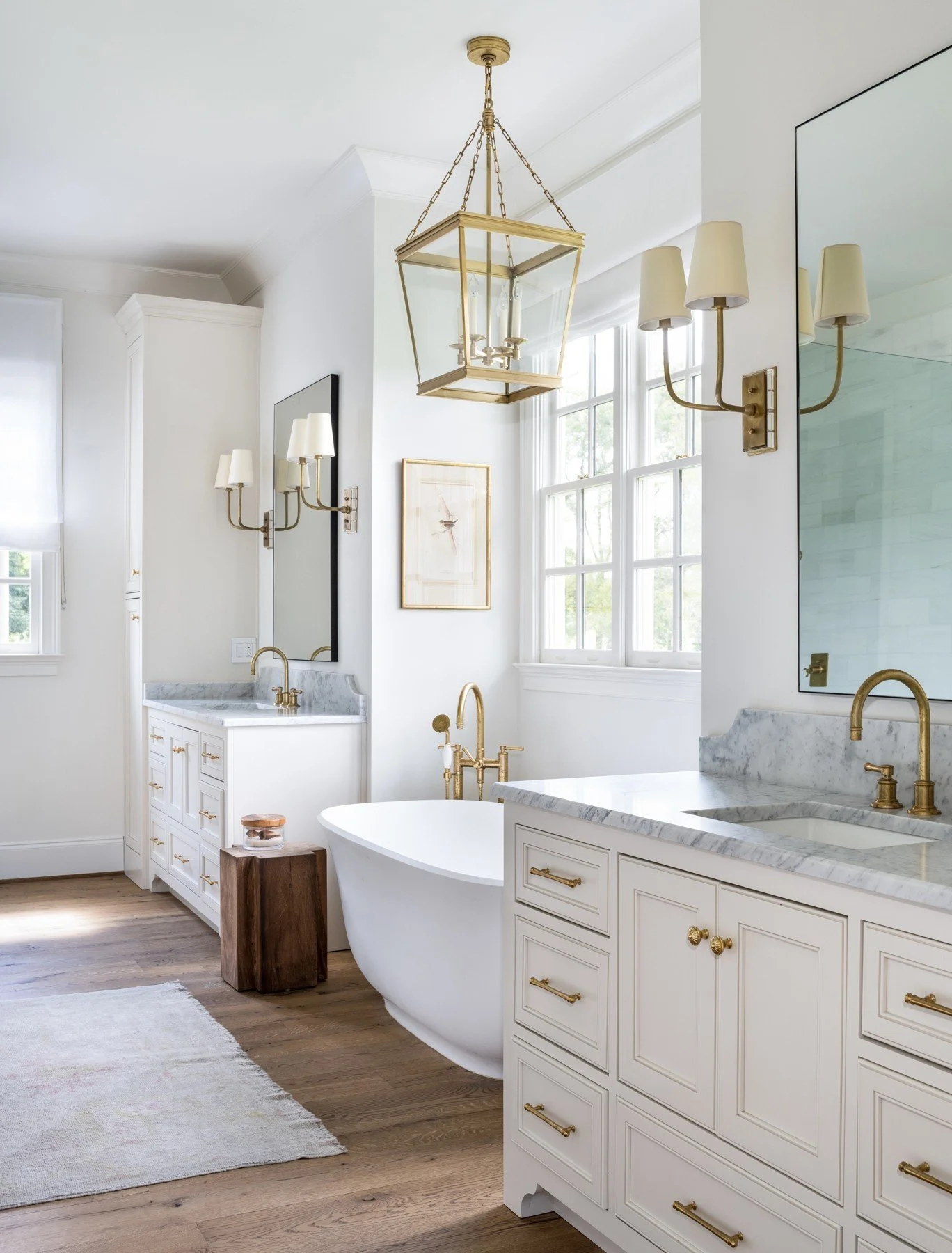 A bright bathroom with white walls, gold fixtures, a freestanding bathtub, marble countertops, and a gold chandelier hanging from the ceiling, along with wall-mounted lights, large windows with white trim, and wooden flooring.
