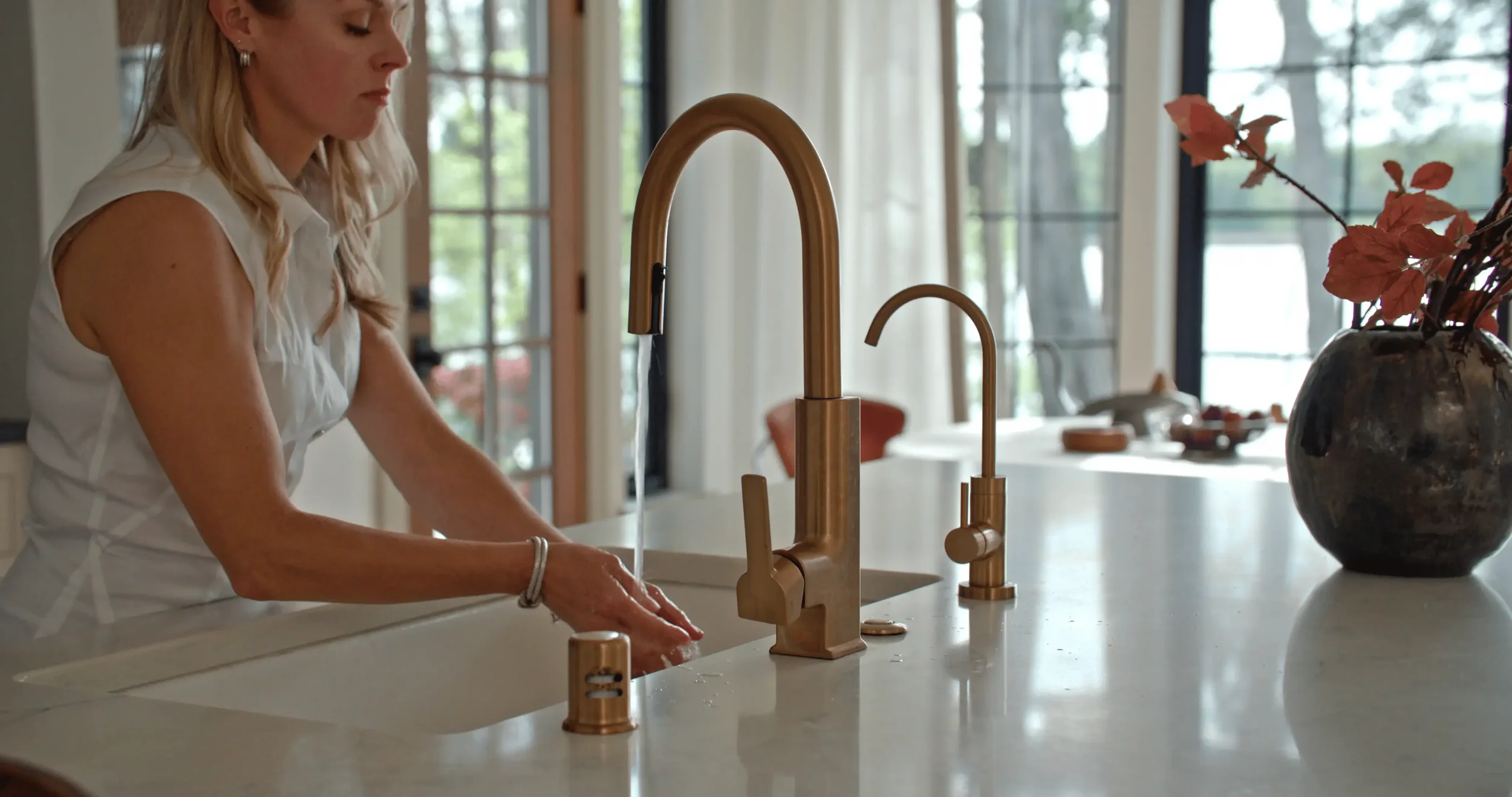 Woman washing her hands at a modern gold kitchen faucet beside a black vase of red branches