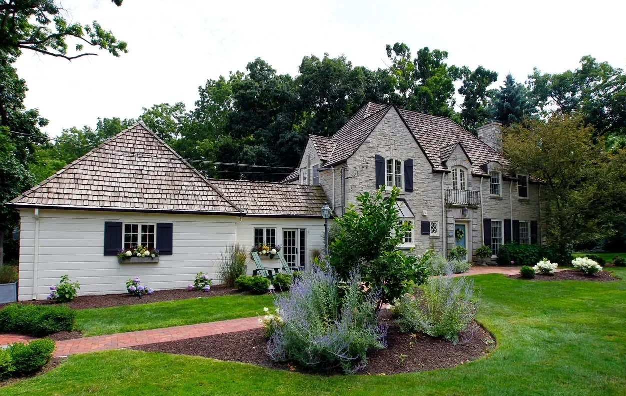 A large house with a stone and white siding exterior surrounded by a well-maintained lawn and garden with bushes and flowers.