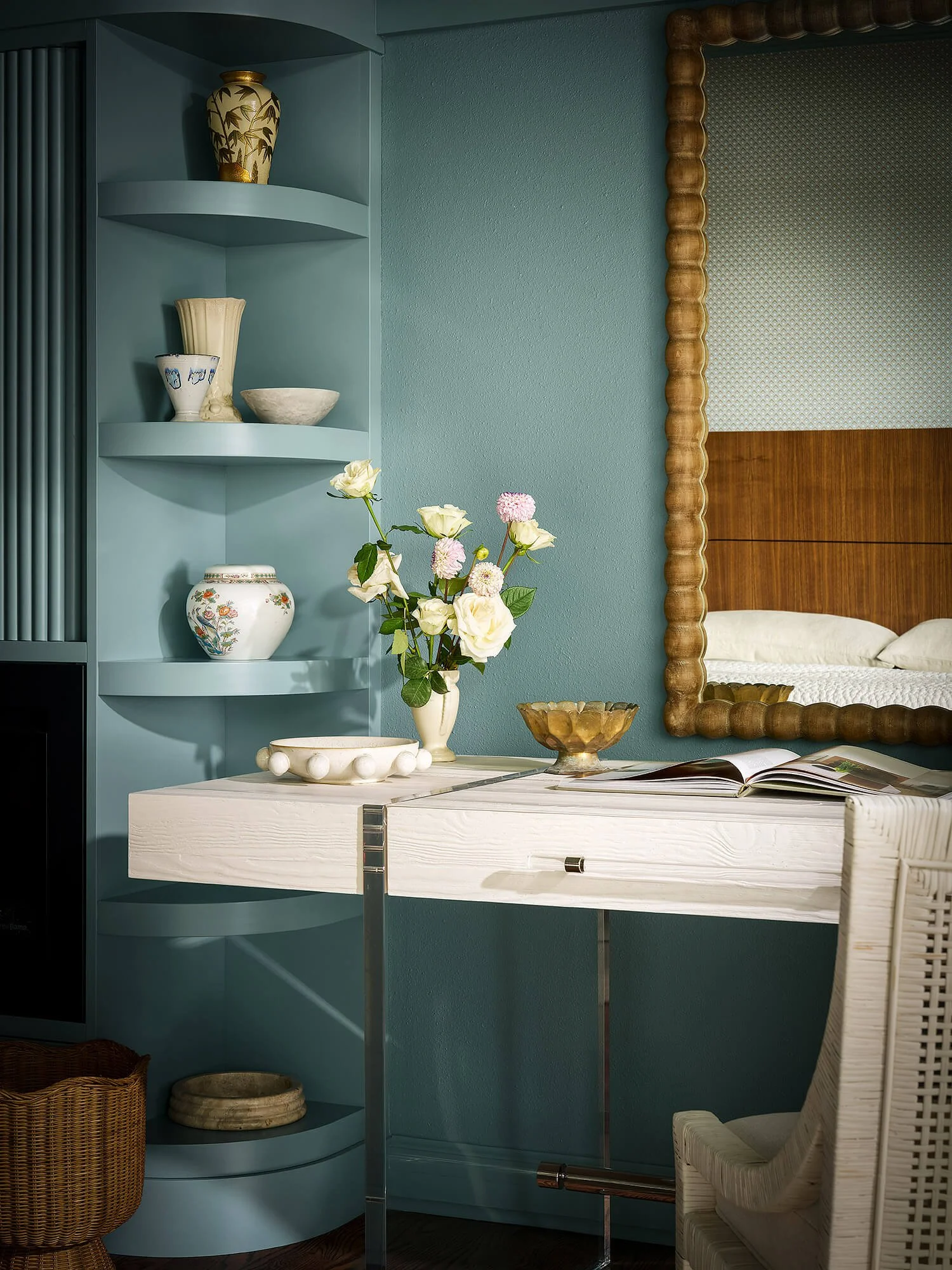 A side view of a corner of a room with a pale blue wall, a light-colored wood table with a glass of water on top, a white wicker chair with a cushion, and a blue built-in shelving unit holding decorative vases, bowls, and a floral arrangement.