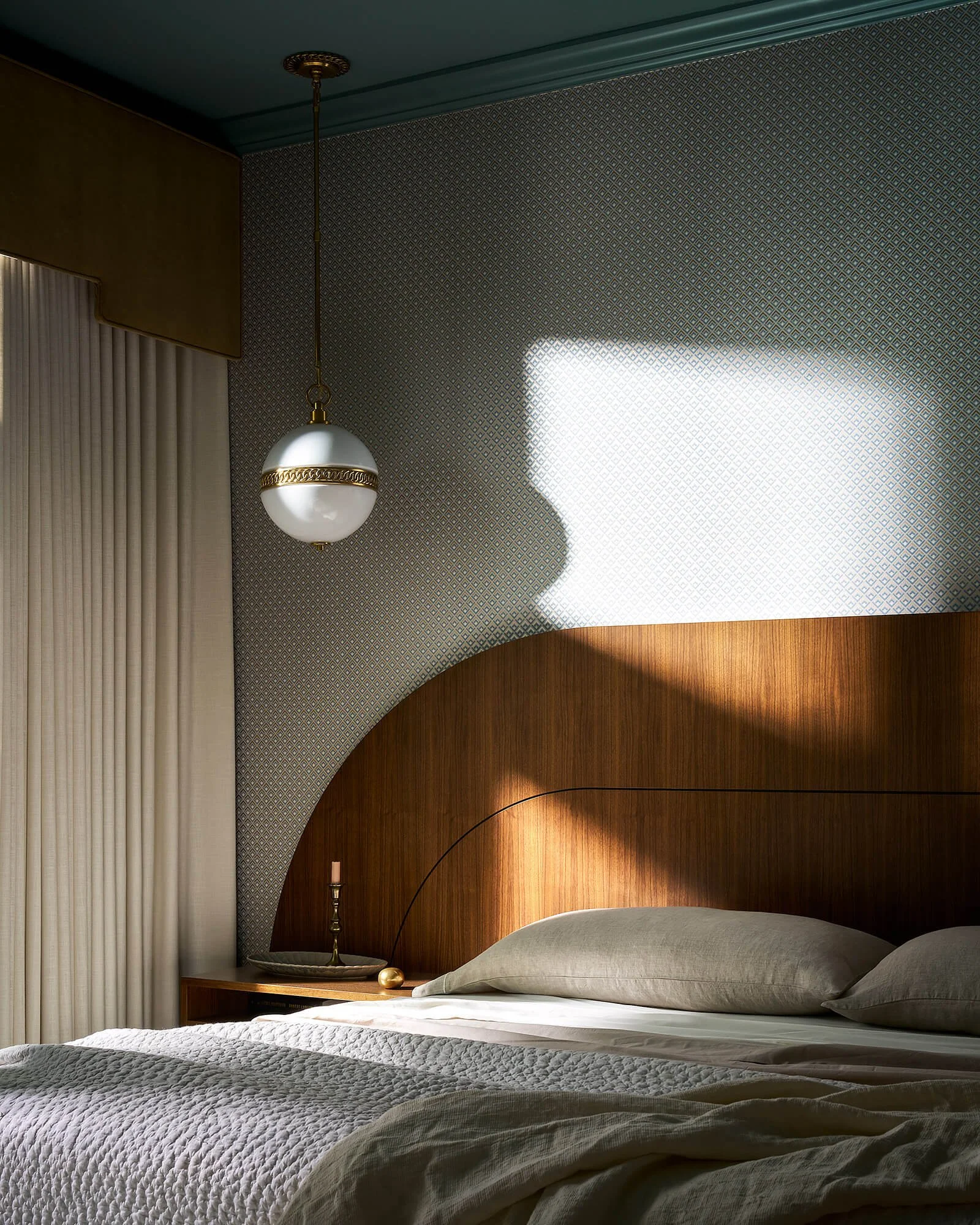 Bedroom with a bed, grey quilted blanket, pillows, a wooden headboard, a nightstand, and a lamp, with patterned wallpaper and curtains, lit by natural light.