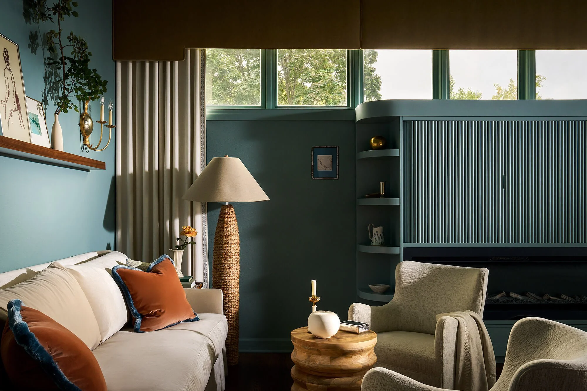 Elegant teal living room with a white sofa, orange accent pillows, woven floor lamp, armchairs, round wood coffee table, fireplace, and large windows