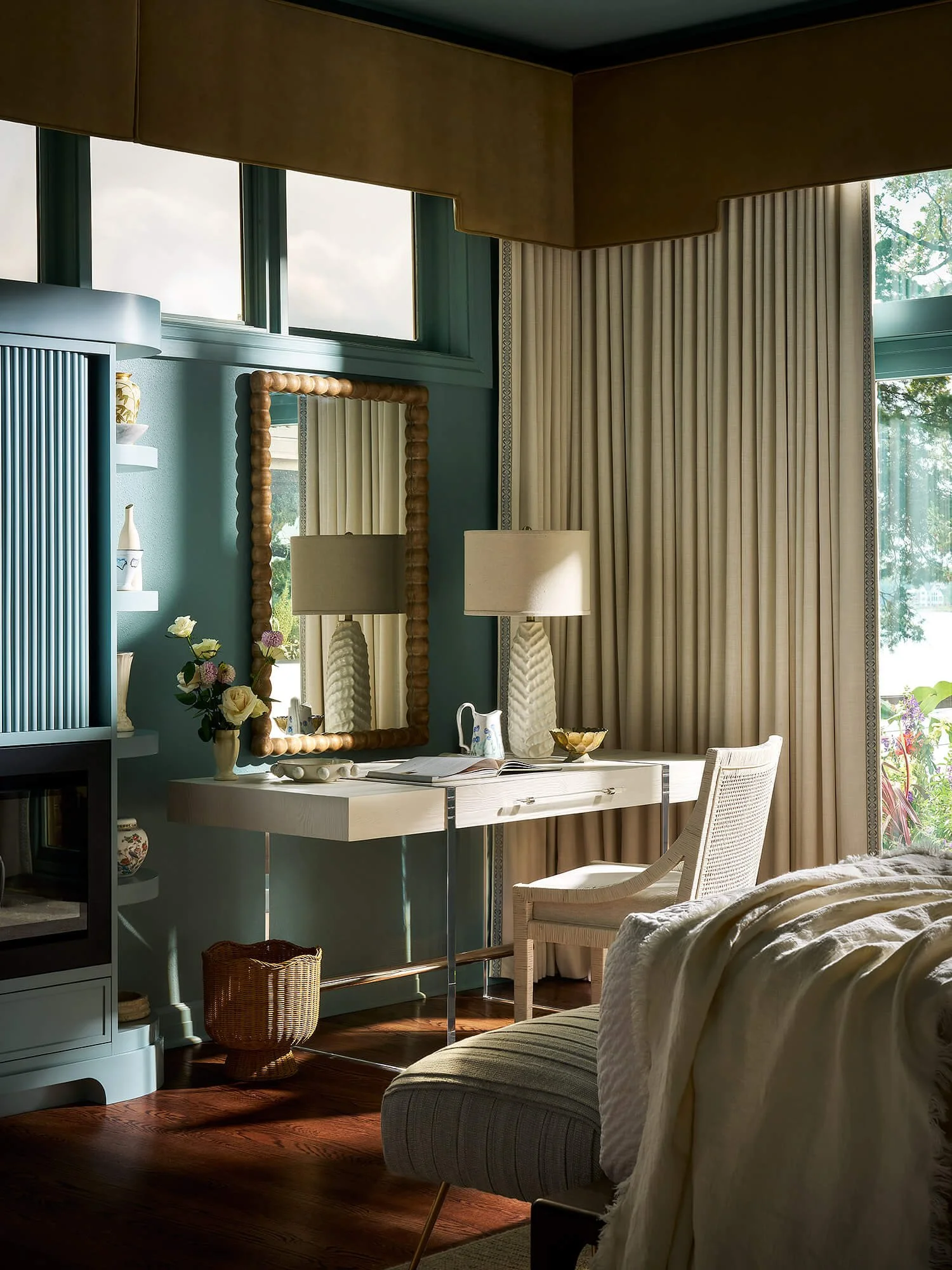 A cozy living room corner with beige curtains, a white desk, a mirror with a wooden frame, a table lamp, and some decorative items on the desk, with natural light coming through large windows.