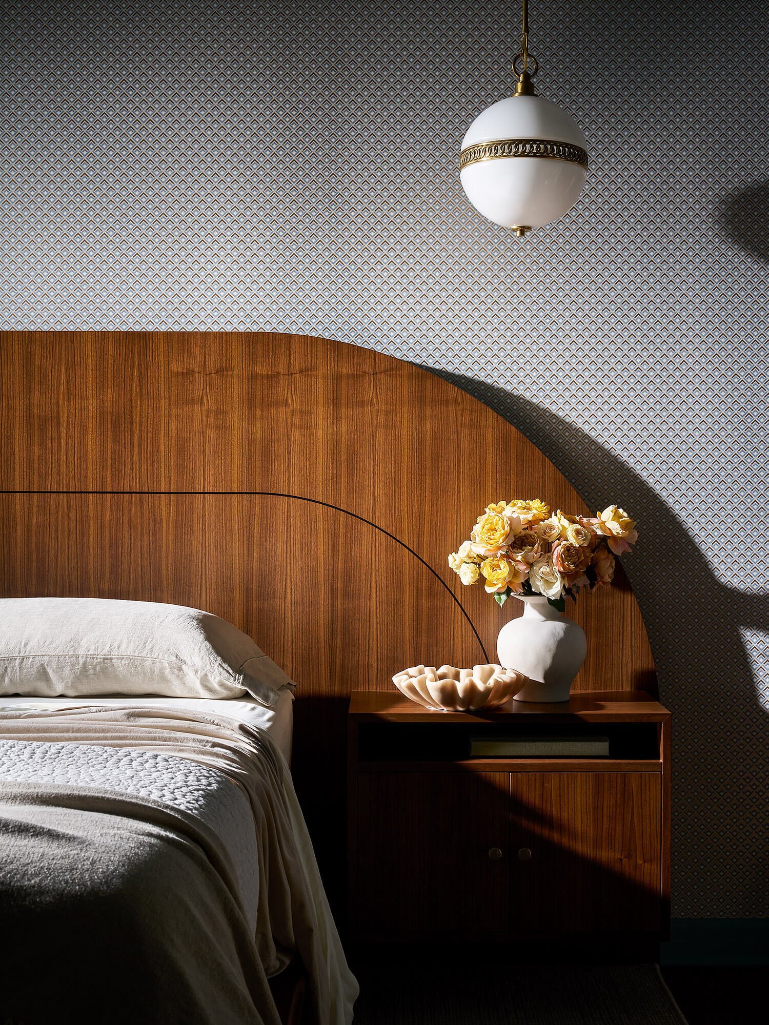 A bedroom with a bed covered in white bedding, a wooden headboard, and a dark wooden nightstand holding a white vase with pink and cream roses, a white decorative bowl, and a patterned wallpaper with a hanging white and gold lamp.