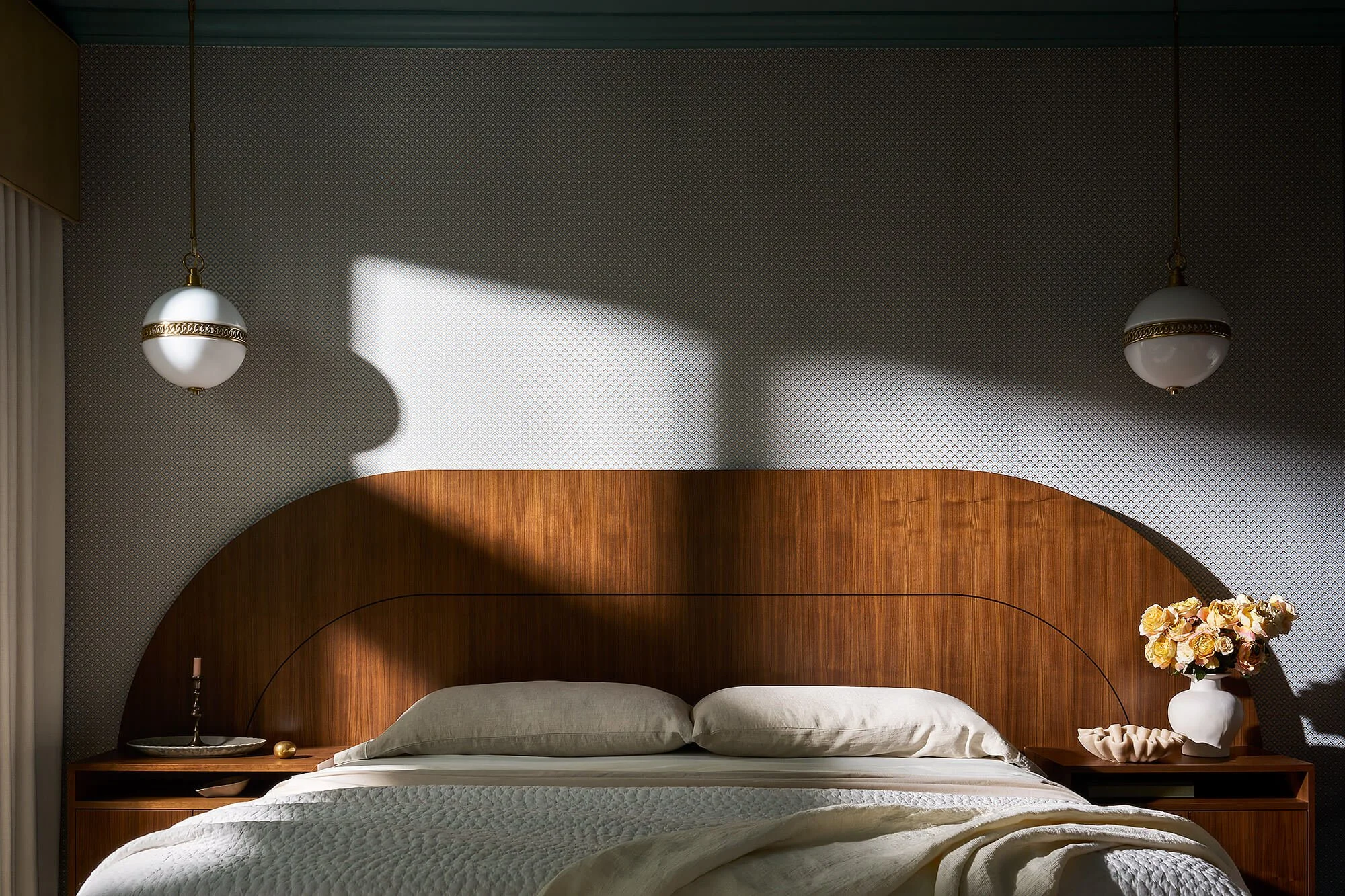 A cozy bedroom with a wooden headboard, bedside lamps, a window with curtains, a vase of flowers, and a cozy blanket on the bed.
