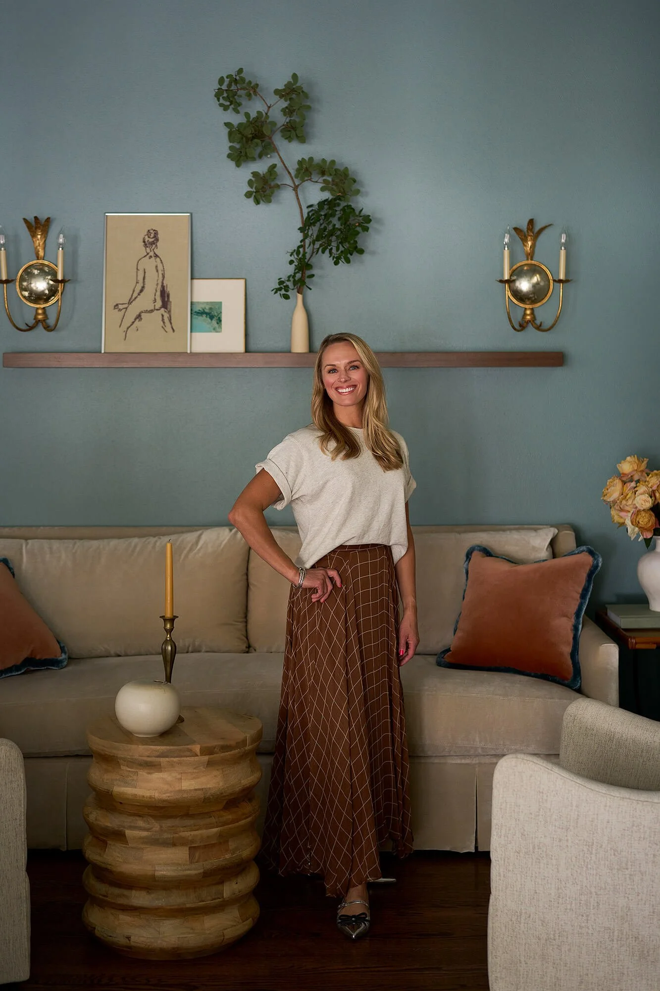 Emily Frank, founder and principal of Frank & Co.  Emily with long blonde hair, wearing a beige top and long brown checkered skirt, stands in a living room with a beige sofa, decorative pillows, a candle on a round wooden table, and framed artwork.