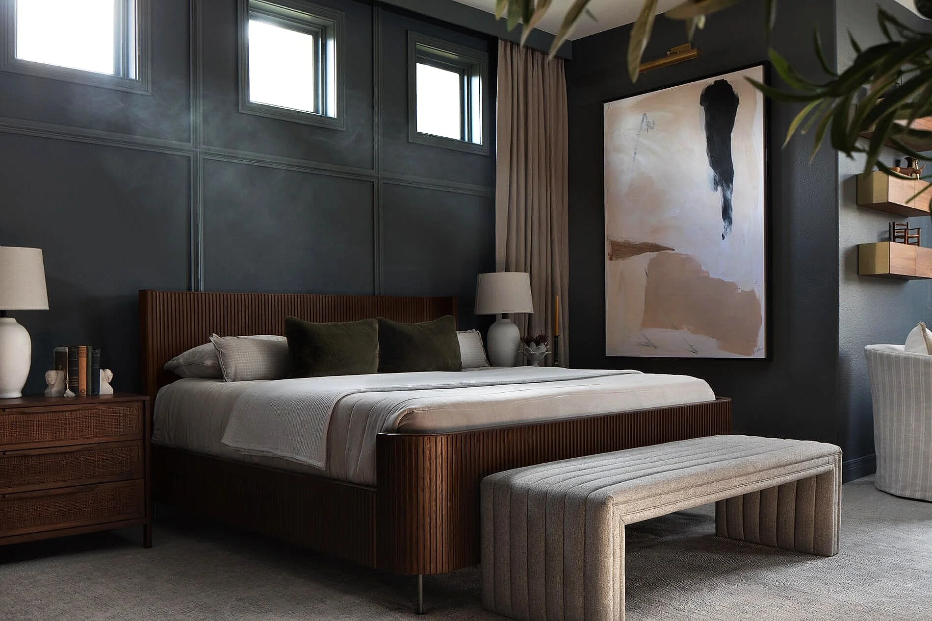 Modern dark bedroom with a wood slatted bed, white bedding, abstract wall art, bedside tables, and upholstered bench