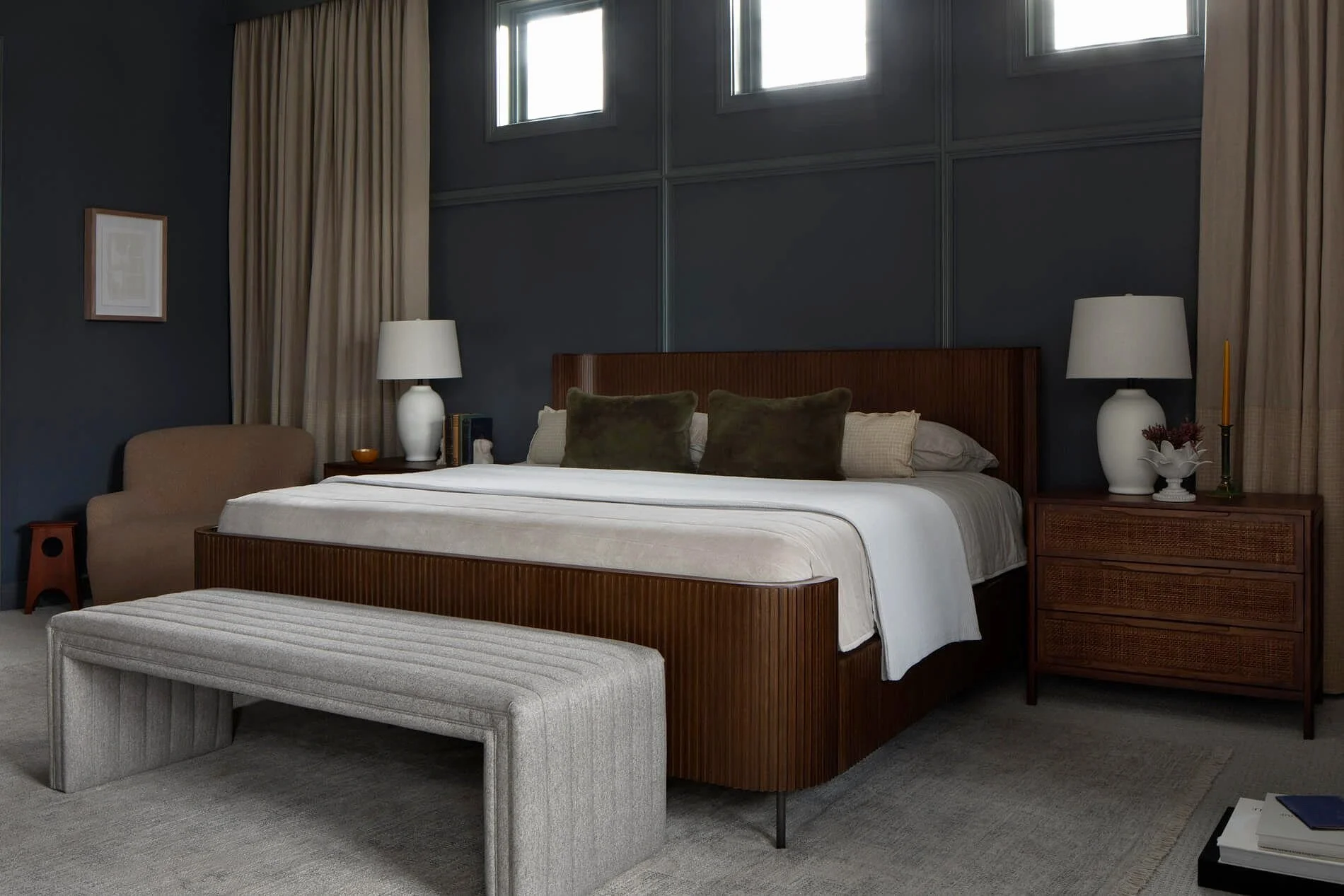 A modern bedroom with dark blue walls and framed artwork, featuring a large wooden bed with white bedding and pillows, matching bedside tables with lamps, and a textured ottoman at the foot of the bed.