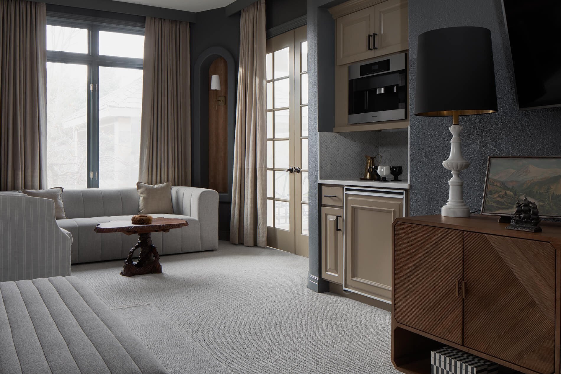 A cozy living room with a large window and beige curtains, a white sofa with beige pillows, a small wooden coffee table, and a side cabinet with a table lamp, painting, and decorative items.