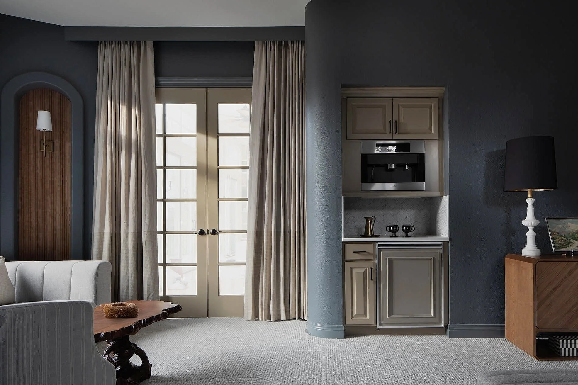 Interior view of a living room with tall glass doors with beige curtains, a white sofa, a wooden coffee table, a built-in beige cabinet with a coffee machine and decorative items, a black lampshade on a white lamp, and dark gray walls.