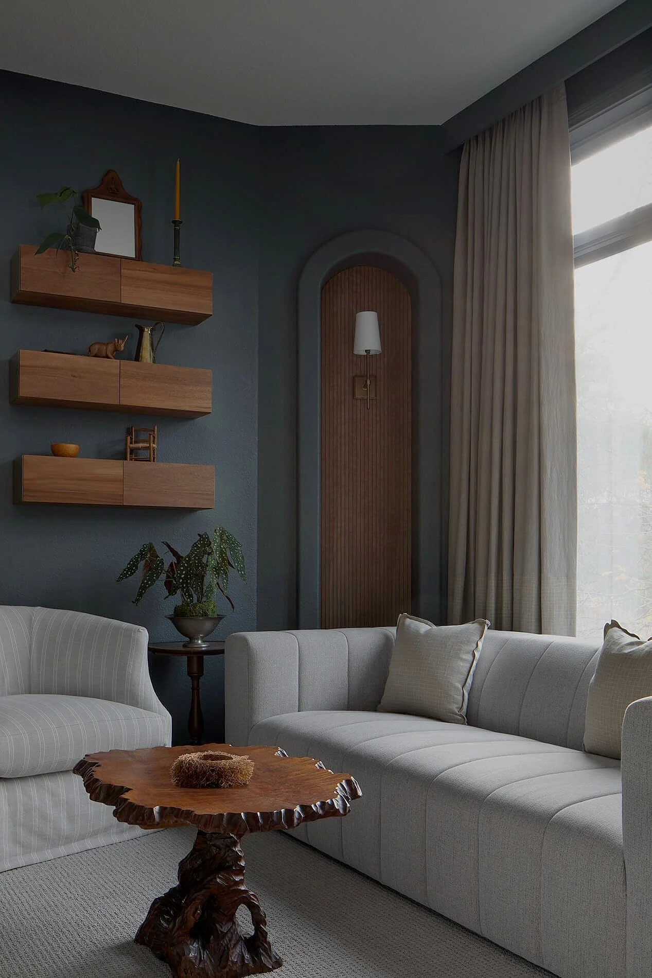 Living room with white sofa, wooden coffee table, and wall-mounted shelves with decor, cream curtains, and a window letting in natural light.