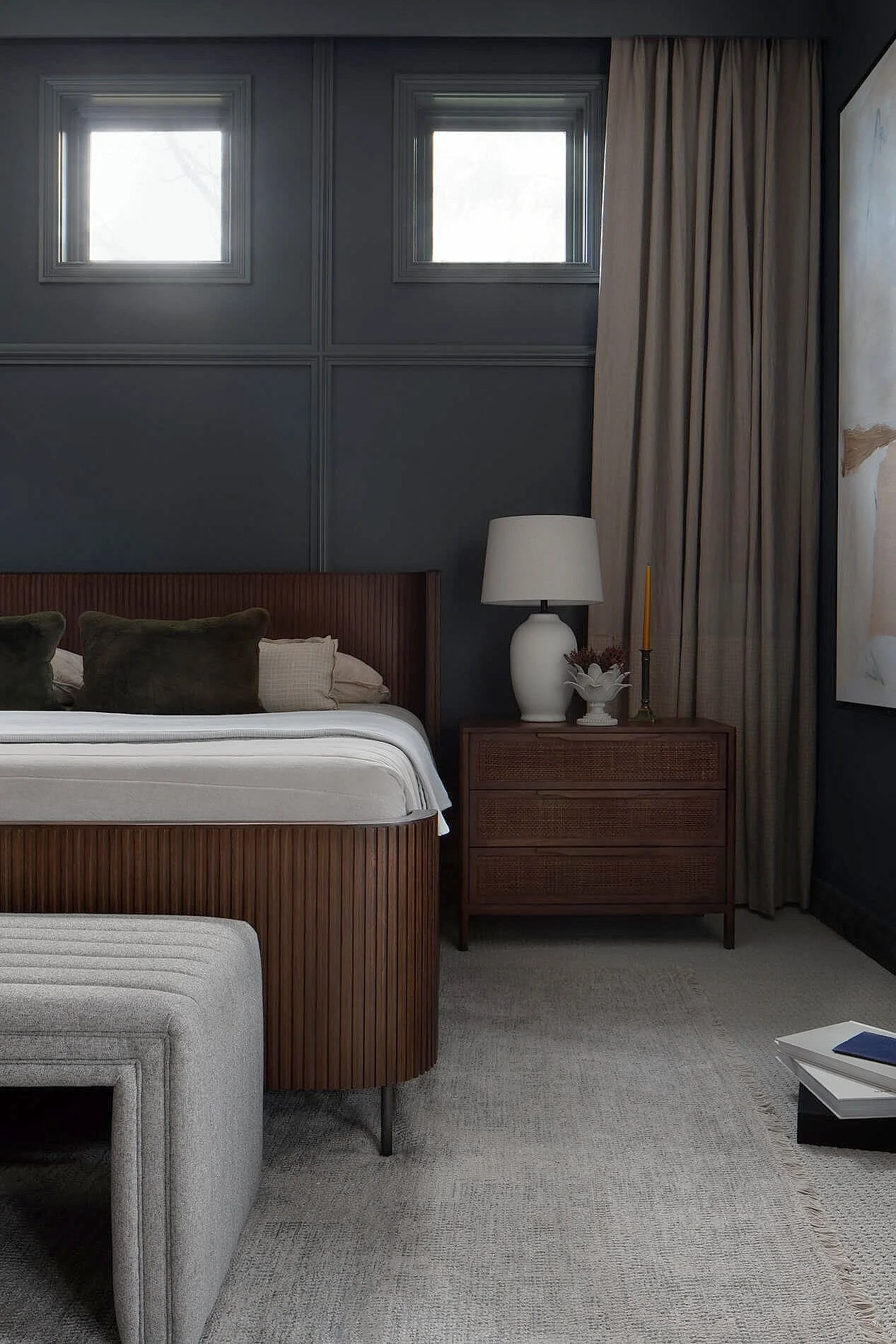 A bedroom with two small high windows, dark blue paneled walls, a wooden bed with a white mattress, green pillows, and beige curtains. There is a wooden nightstand with a white lamp, a decorative bowl, and a tall candle, plus a framed artwork on the wall.