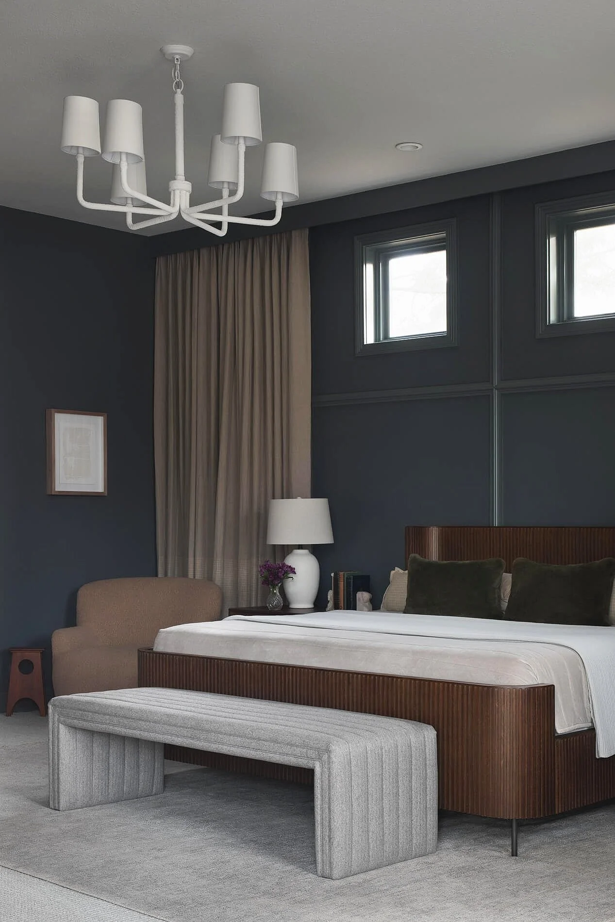 A bedroom with dark walls, a white chandelier, two windows, beige curtains, a bed with a wooden headboard, pillows, a white table with a white lamp and purple flowers, a beige armchair, a gray bench, and a white ottoman.