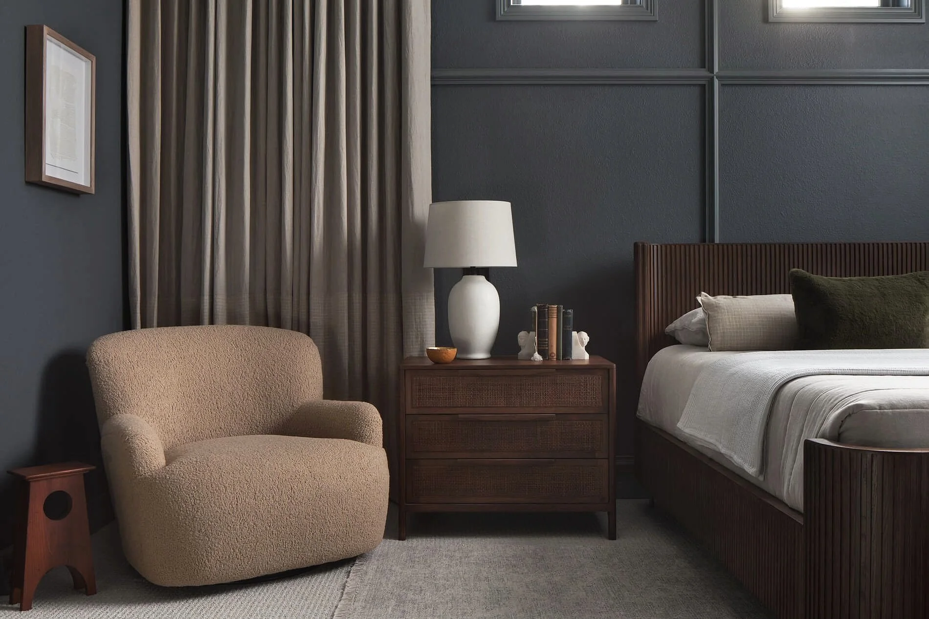 A cozy bedroom corner with a beige, plush armchair, a wooden bedside table with a large white table lamp, a few books, and decorative items. The bed has a wooden frame with a striped blanket, and there are two pillows, one dark green and the other beige. The wall behind has dark gray paneling, and to the left, there are beige curtains covering a window. A framed picture hangs on the dark wall.