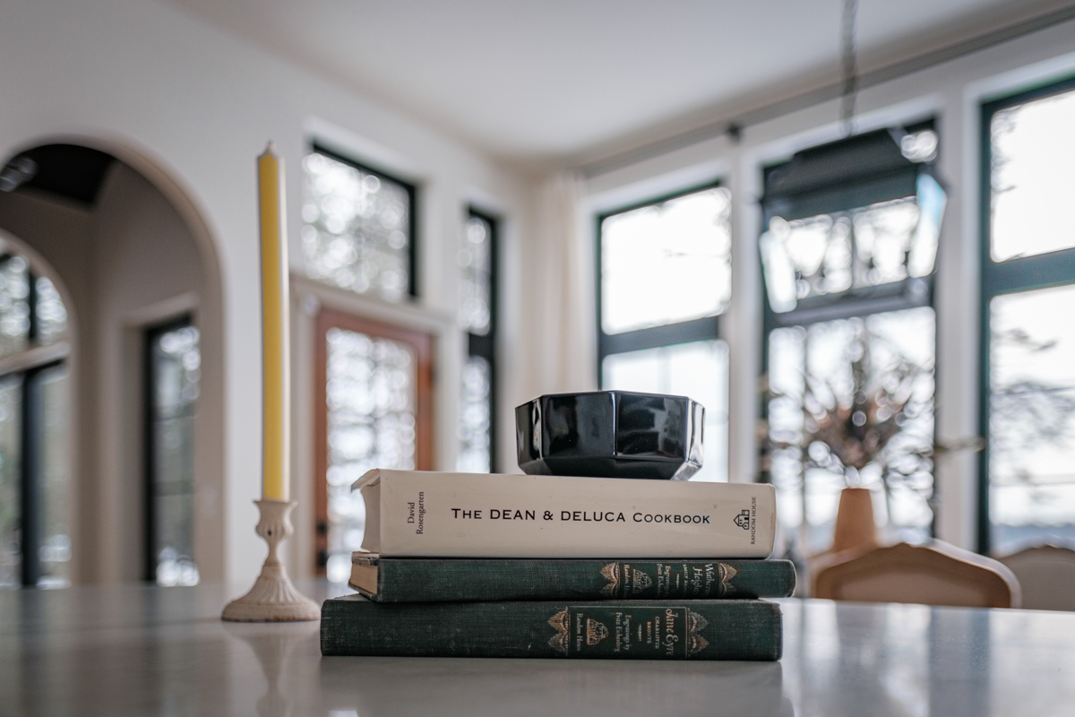 Stack of three books on a table with a black bowl on top, a yellow candle in a white holder to the left, and a blurred background of large windows and a plant.