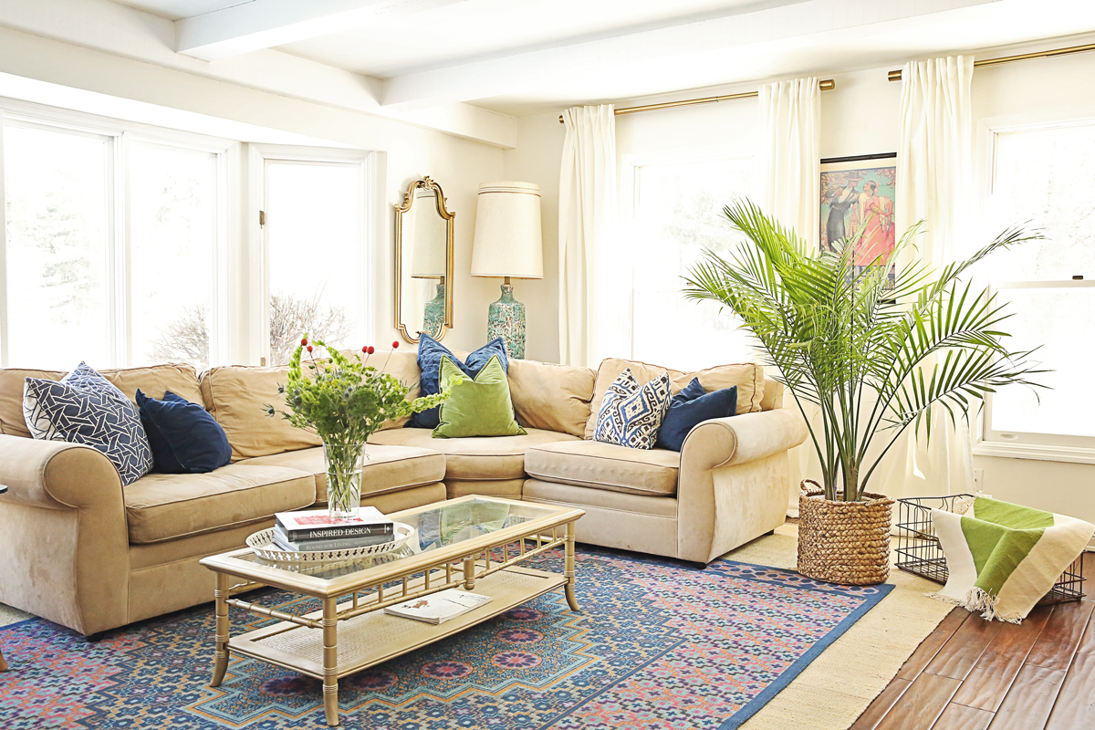 Bright living room with large windows, beige sectional sofa with colorful pillows, glass coffee table with books, green potted plant in a basket, and white curtains.
