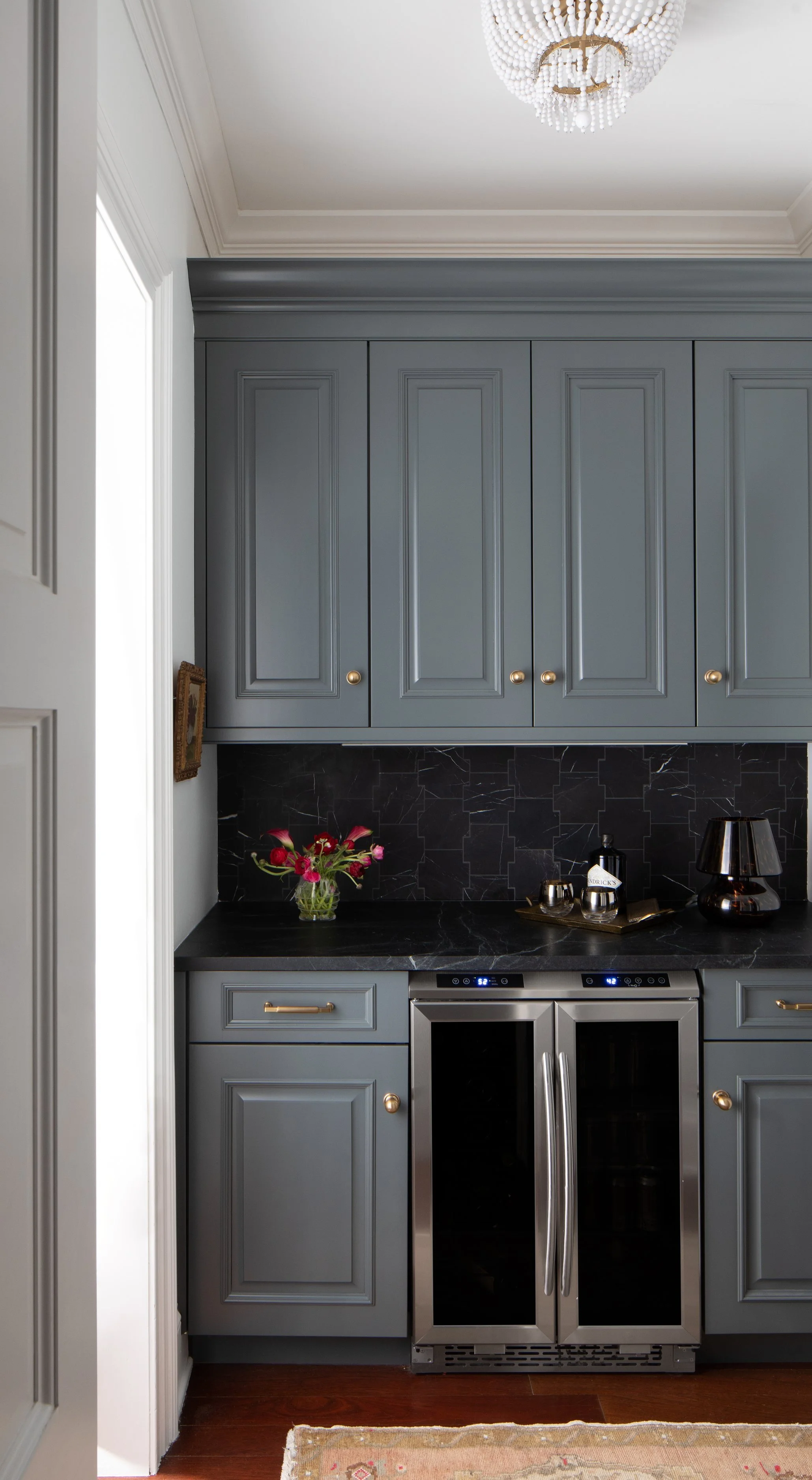 A kitchen with gray cabinets, black marble backsplash, and stainless steel appliances, including a wine cooler. Decor includes a vase with pink flowers, glassware, and a black lamp, with a chandelier hanging from the ceiling.