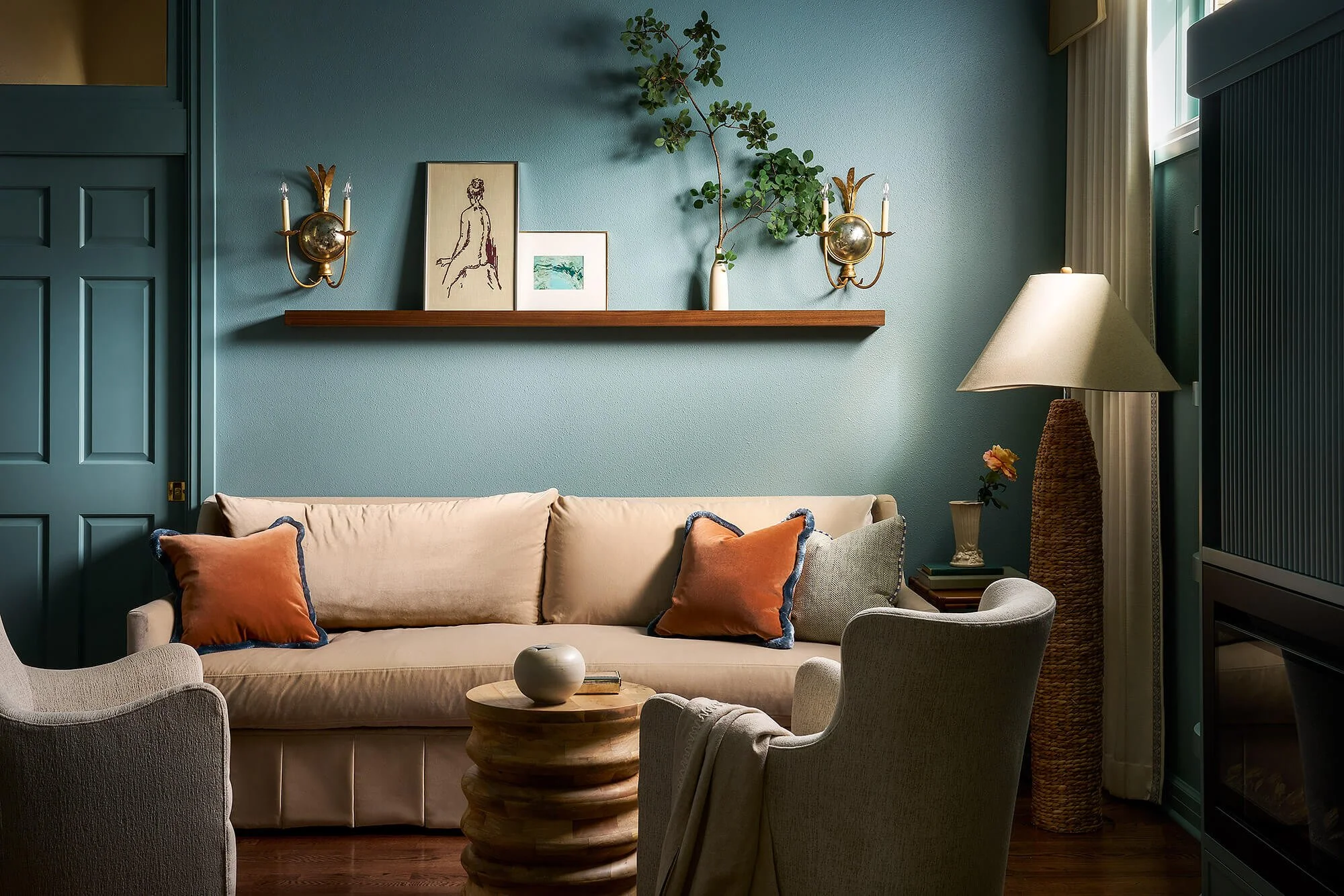 Living room with a beige sofa decorated with orange and blue pillows, a round wooden coffee table, and upholstered chairs. The wall behind the sofa is teal with framed artwork, green plant, and brass wall sconces with candles. A tall woven basket with a plant, a side table with a flower, and a floor lamp are also visible in the room.
