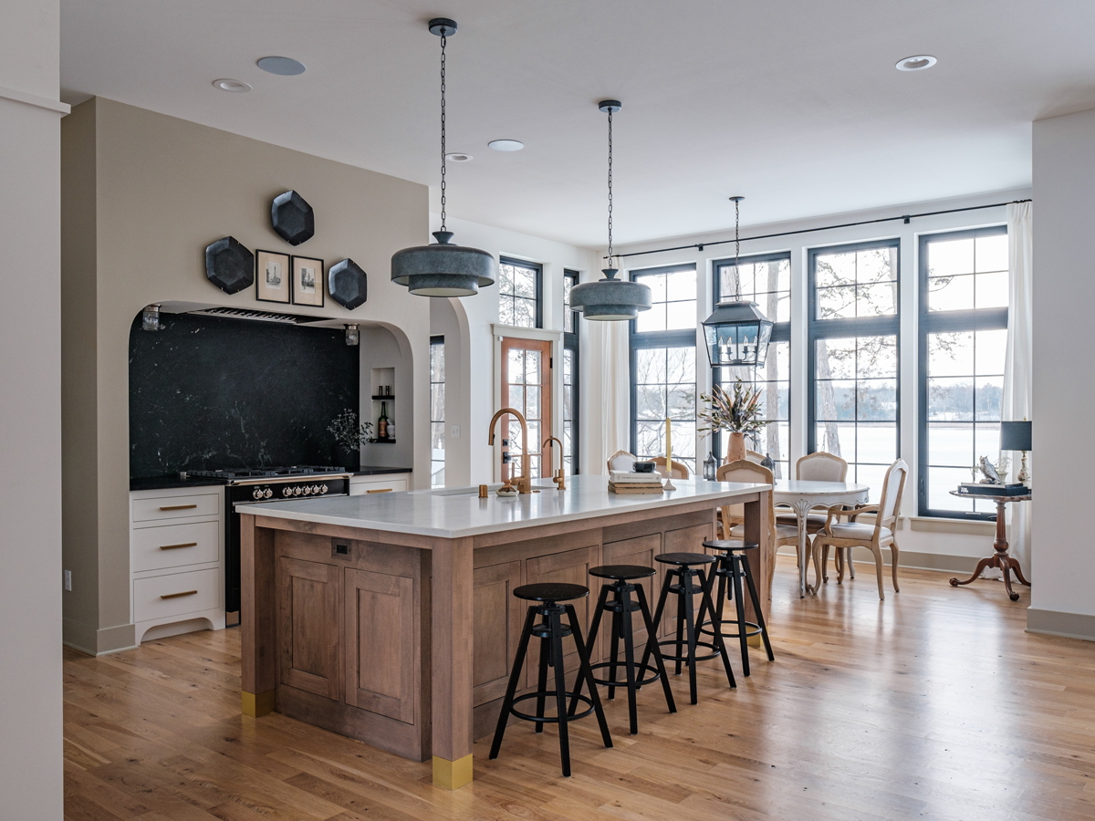 Open-concept kitchen and dining area with a large island, black barstools, pendant lights, and a dining table with chairs by large windows showing an outdoor view.