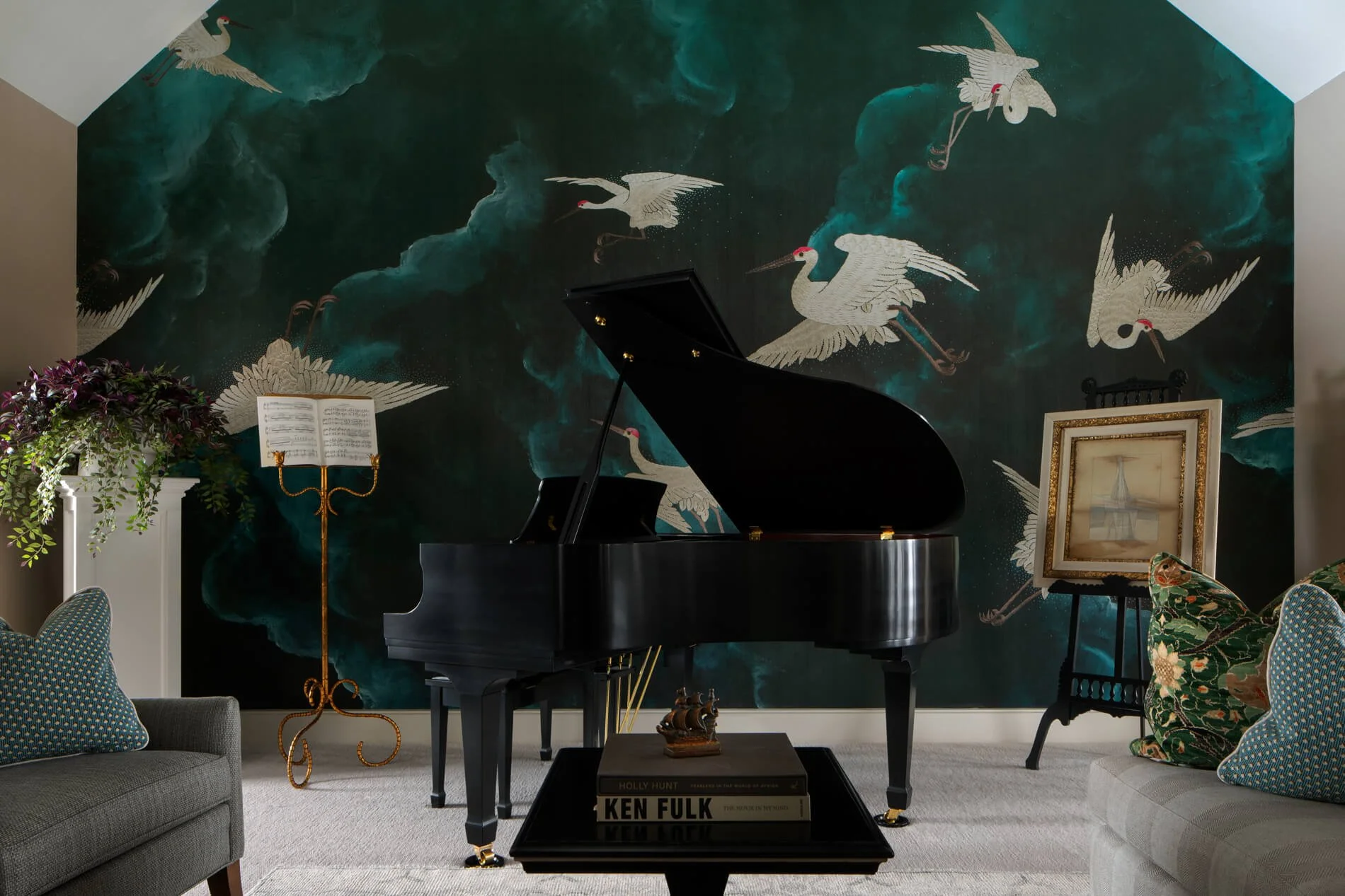 Living room with dark green wall featuring white cranes painted on it, a black grand piano, a stand with sheet music, framed artwork, a small table with books, and various patterned chairs and pillows.