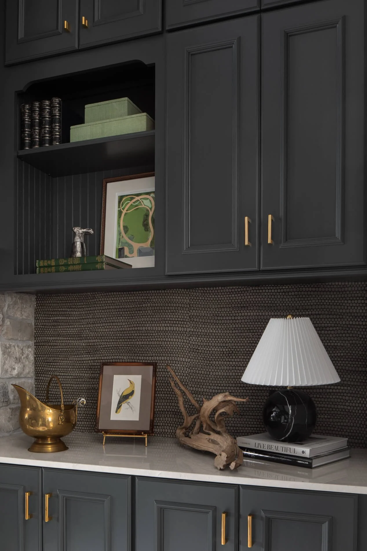 Interior view of a dark gray cabinet with gold handles, decorative objects on the countertop including a gold pitcher, a framed bird illustration, a piece of driftwood, a black and gold table lamp, and books with a stacked magazine titled "Live Beautiful."
