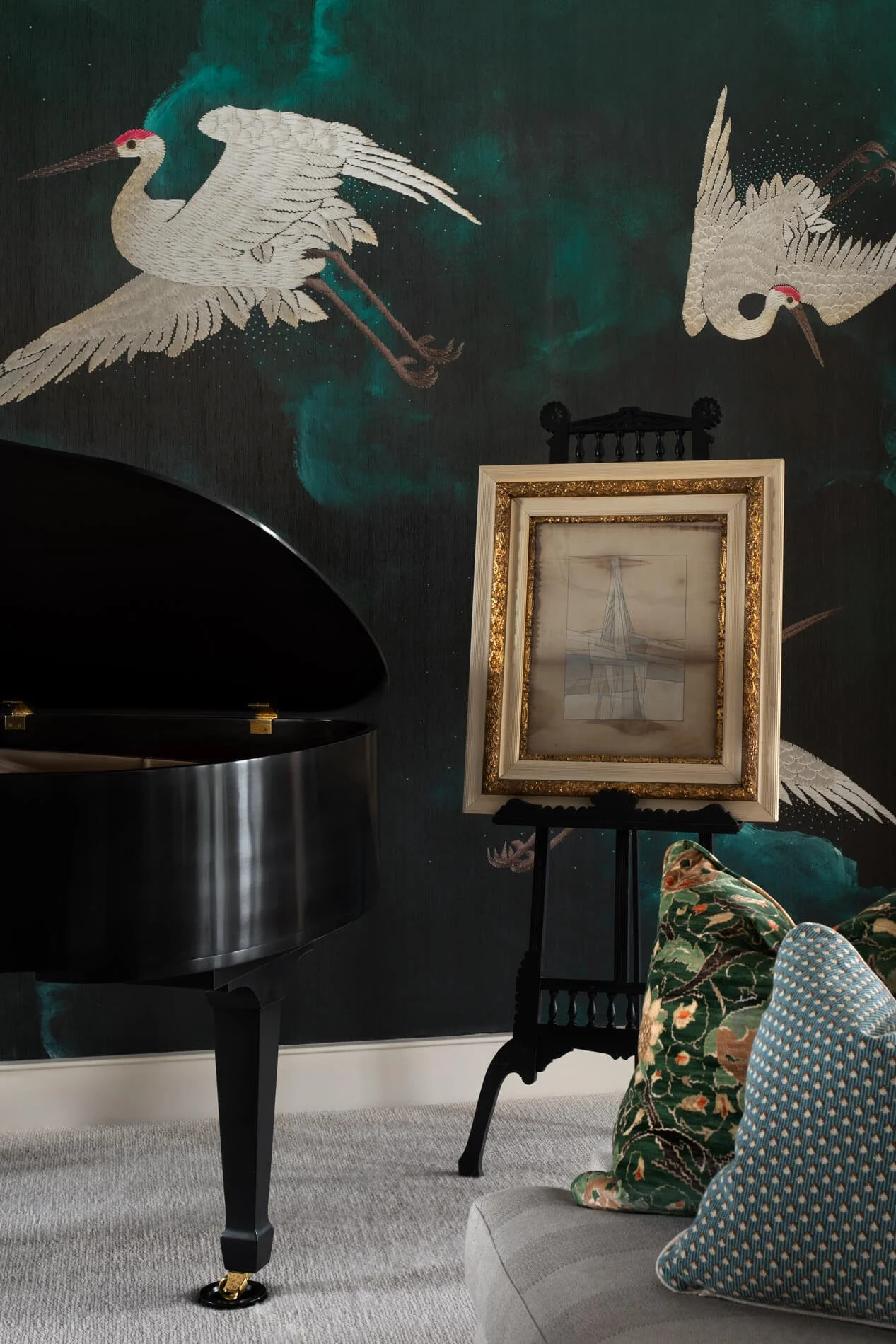 Room with wall art of two white cranes and a framed sketch of a sailing ship, a black grand piano, and patterned cushions on a beige sofa.