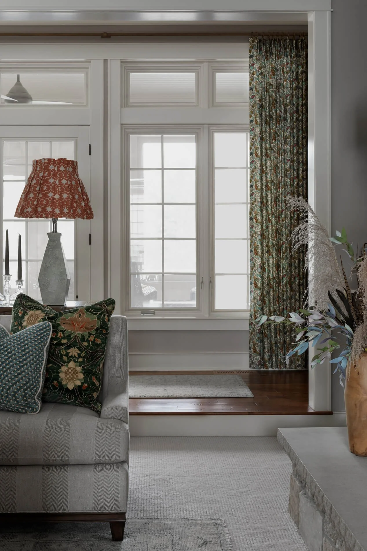 Living room with a beige sofa, patterned pillows, a side table with candlesticks, a lamp with a floral lampshade, large windows with patterned curtains, and decorative plants.